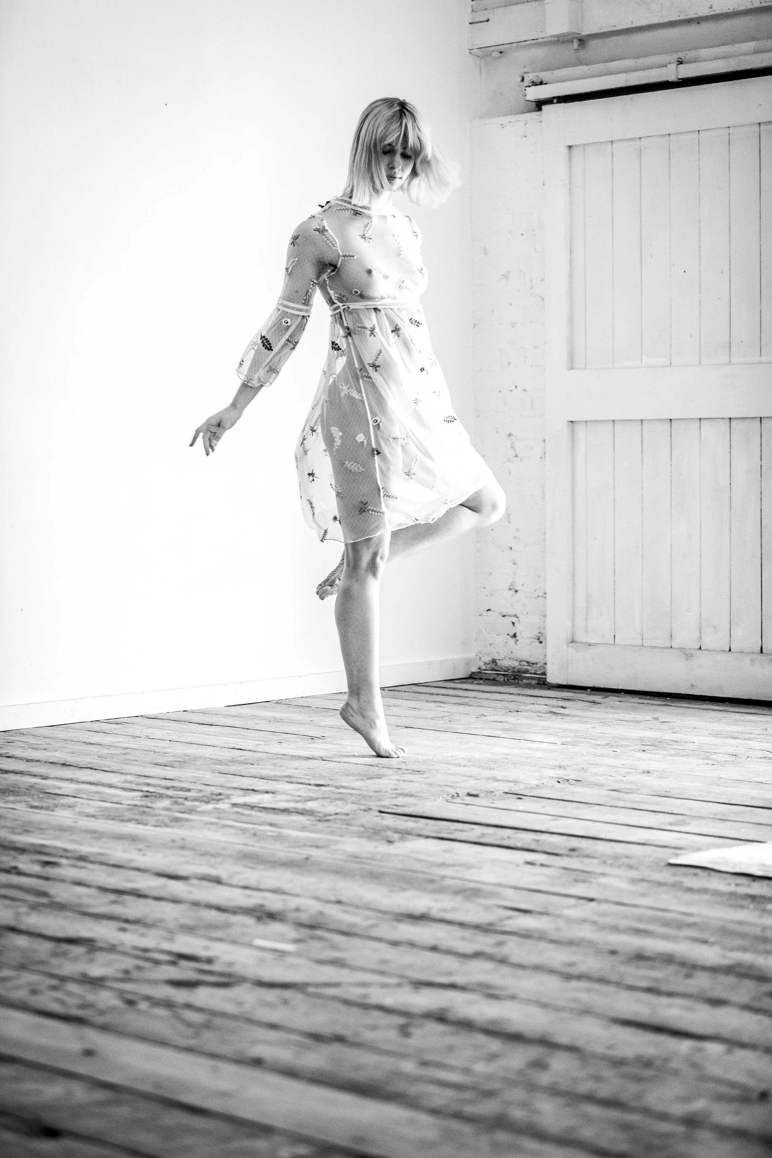 A woman with short hair dancing on a wooden floor in a minimalistic room with white walls and a door.