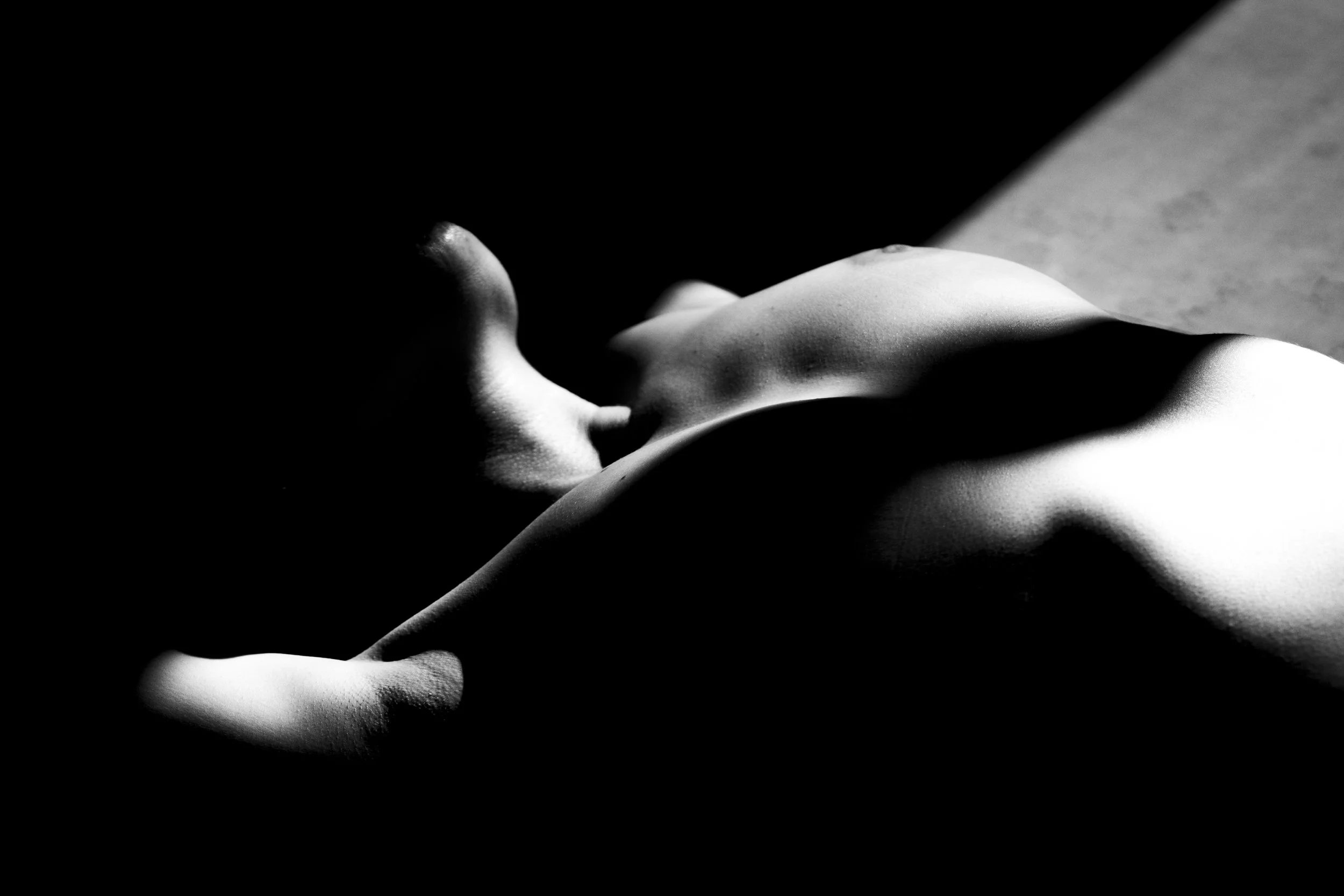 Black and white photo of a topless person lying on their back with their hand covering their face, shadowed parts of their body and background.