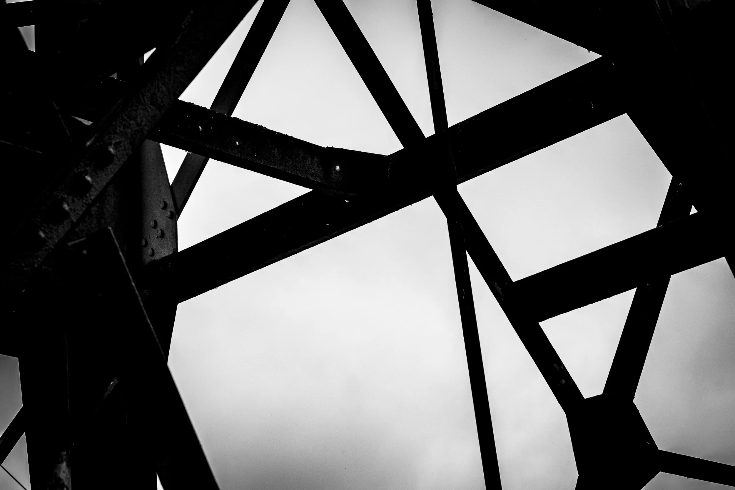 Black and white photo showing the metal framework of a structure, with various beams and a cloudy sky in the background.