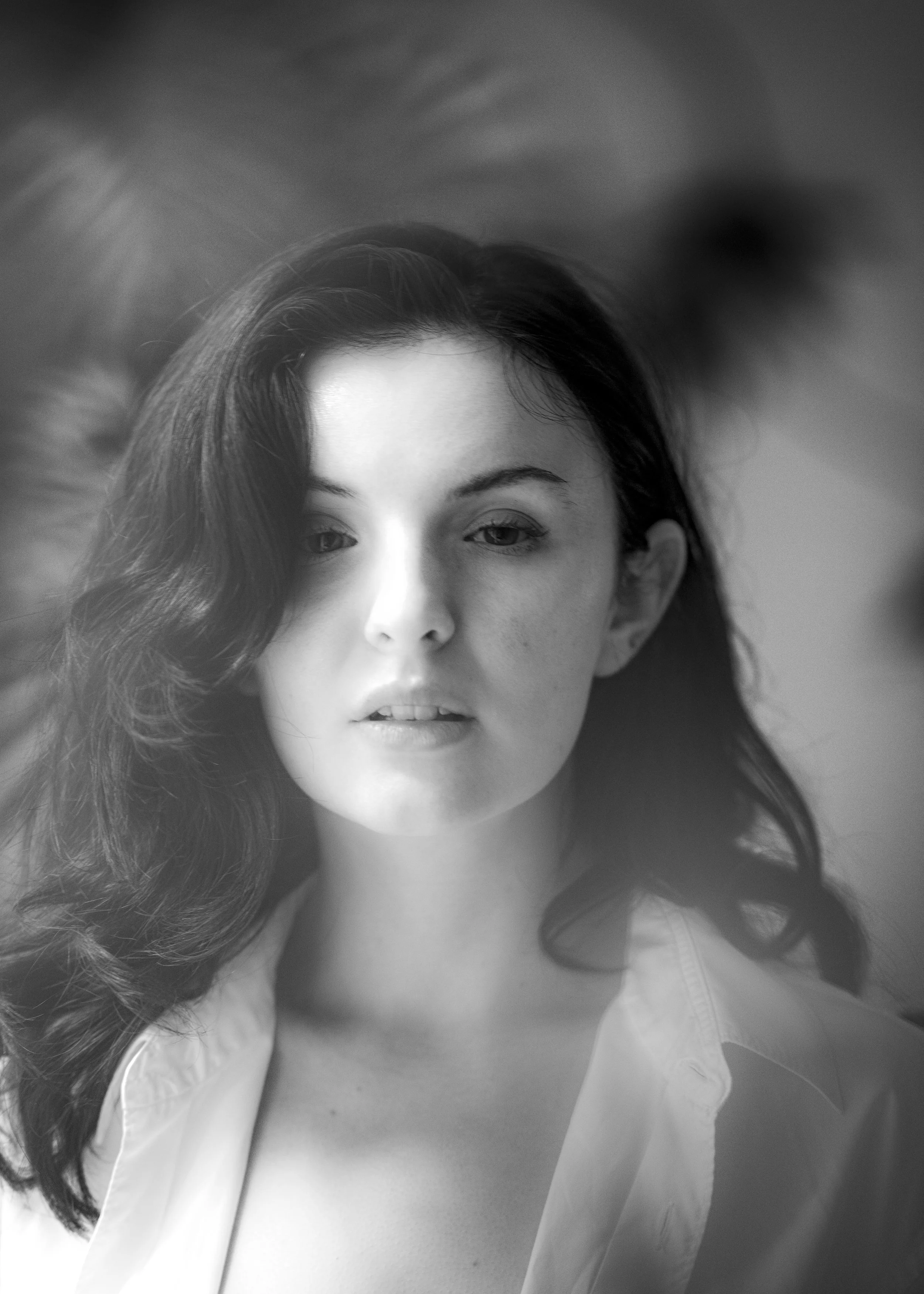 Black and white close-up of a woman's face with wavy hair, wearing a light-colored blouse, looking at the camera with a neutral expression.