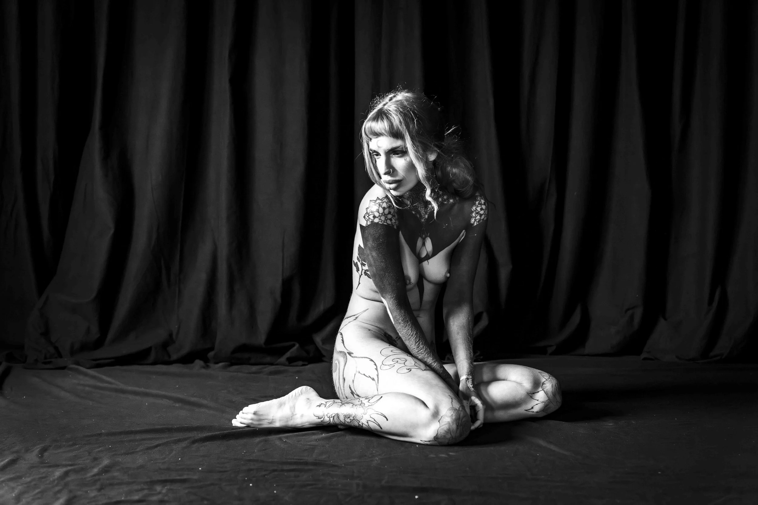 A woman with tattoos sitting on the floor with her legs crossed, facing downward and to the side, in front of black curtains.