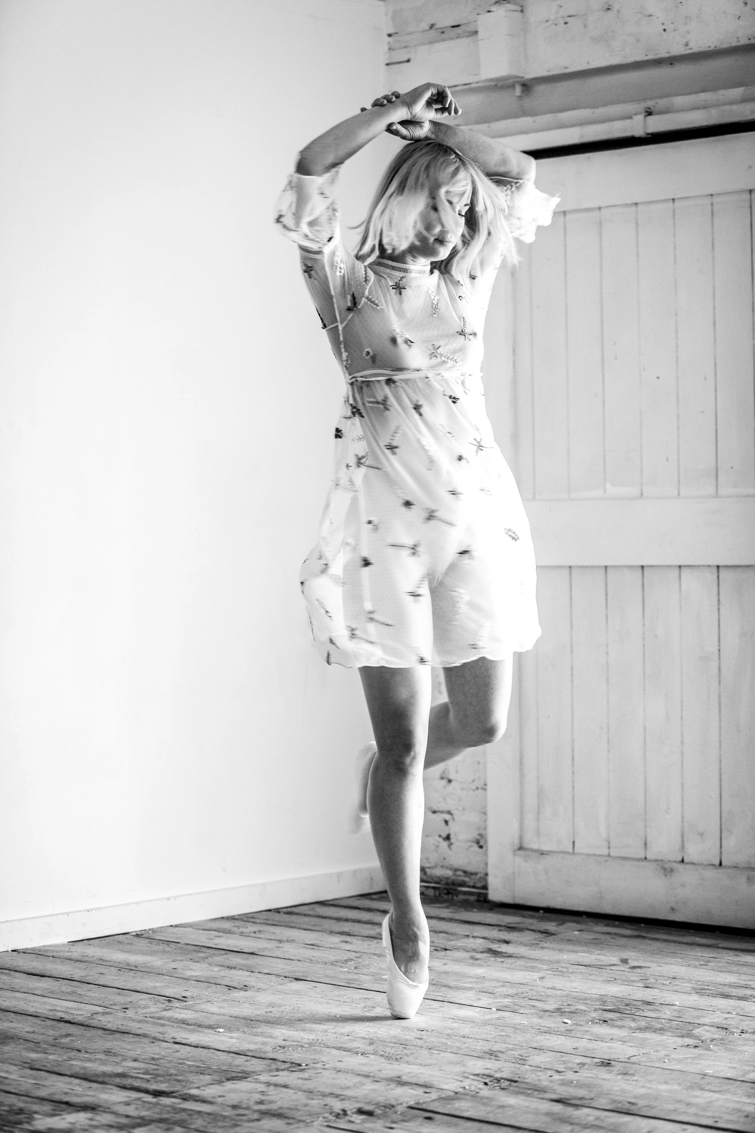A woman in a ballet pose on one leg, with arms raised above her head, standing against a wooden wall and floor in a bright, minimal room.