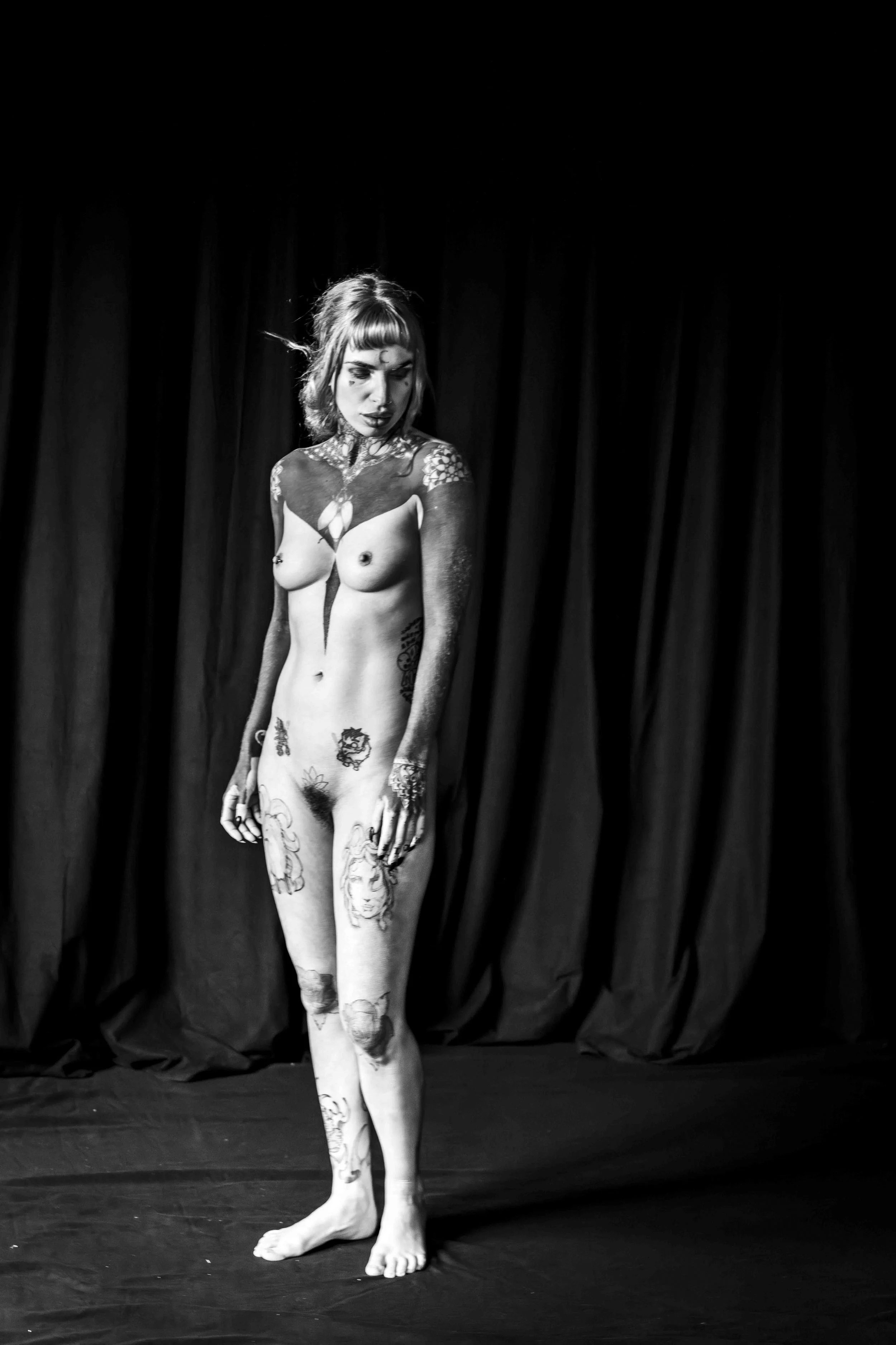A woman with tattoos on her body stands on stage against a dark curtain. She has short hair, tattoos on her arms, torso, and legs, and is looking down with her hands at her sides.