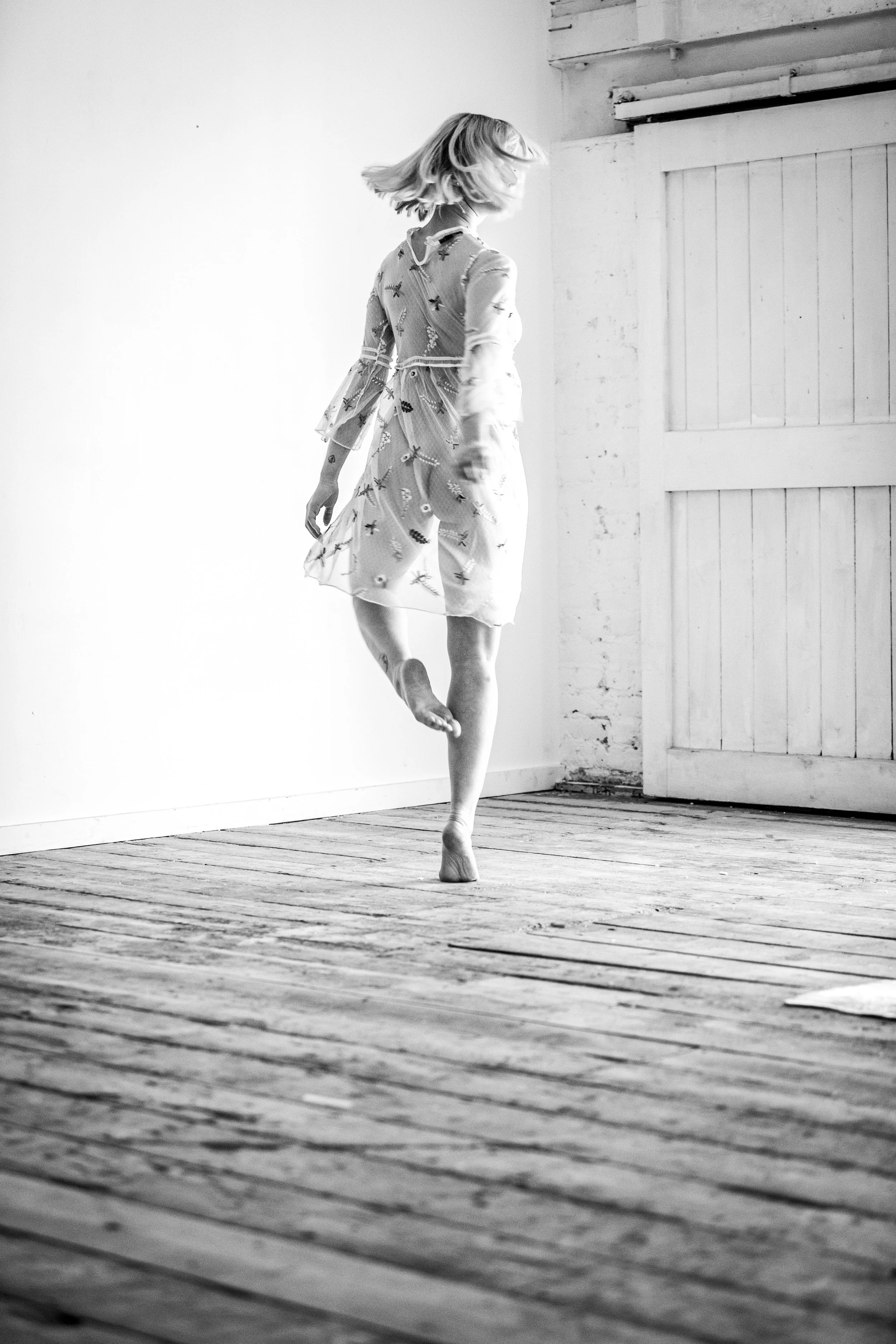A woman dancing on a wooden floor with a bare wall and a door in the background, shown in black and white.