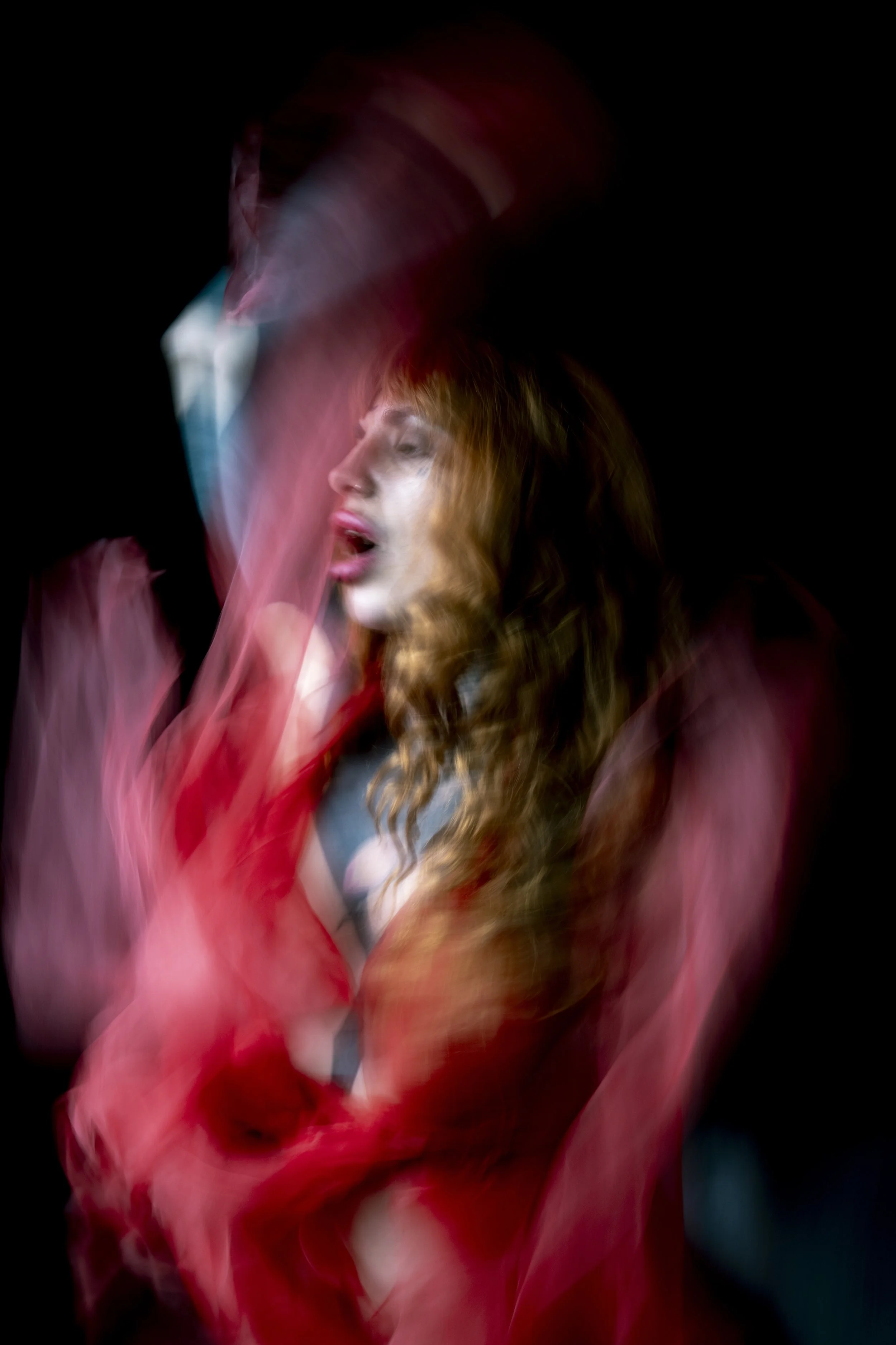 A woman with long, curly red hair and a serene expression, partially obscured by swirling red and pink fabric, against a dark background.
