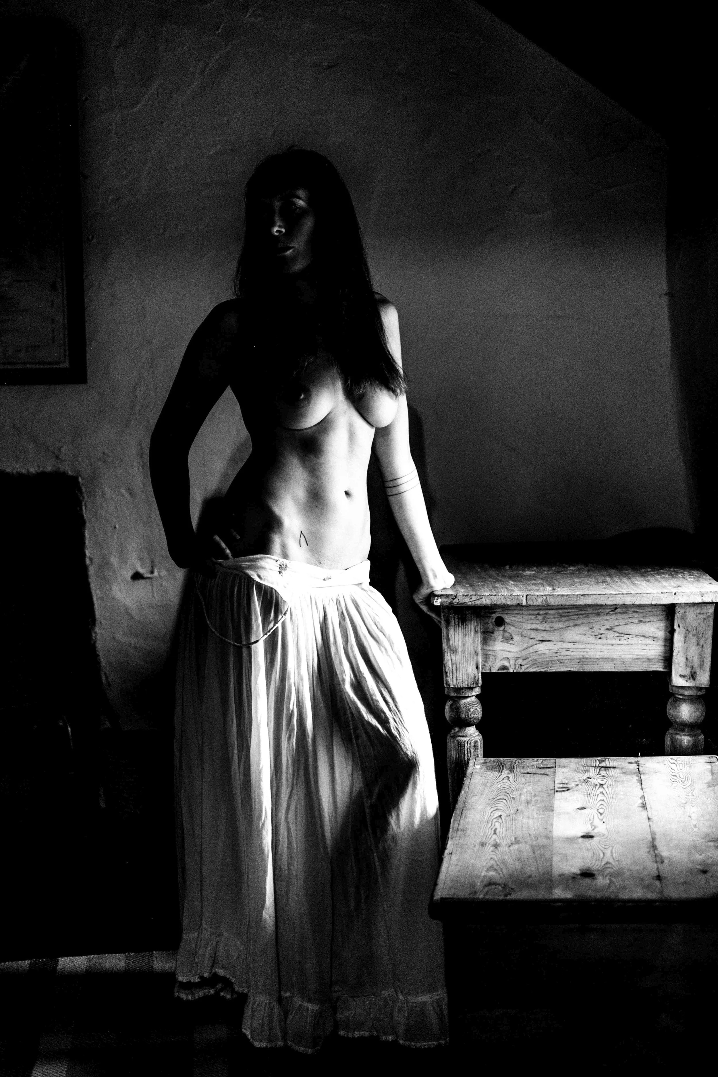 A black and white photo of a woman with long hair, standing next to a wooden table, in a dimly lit room with textured walls.