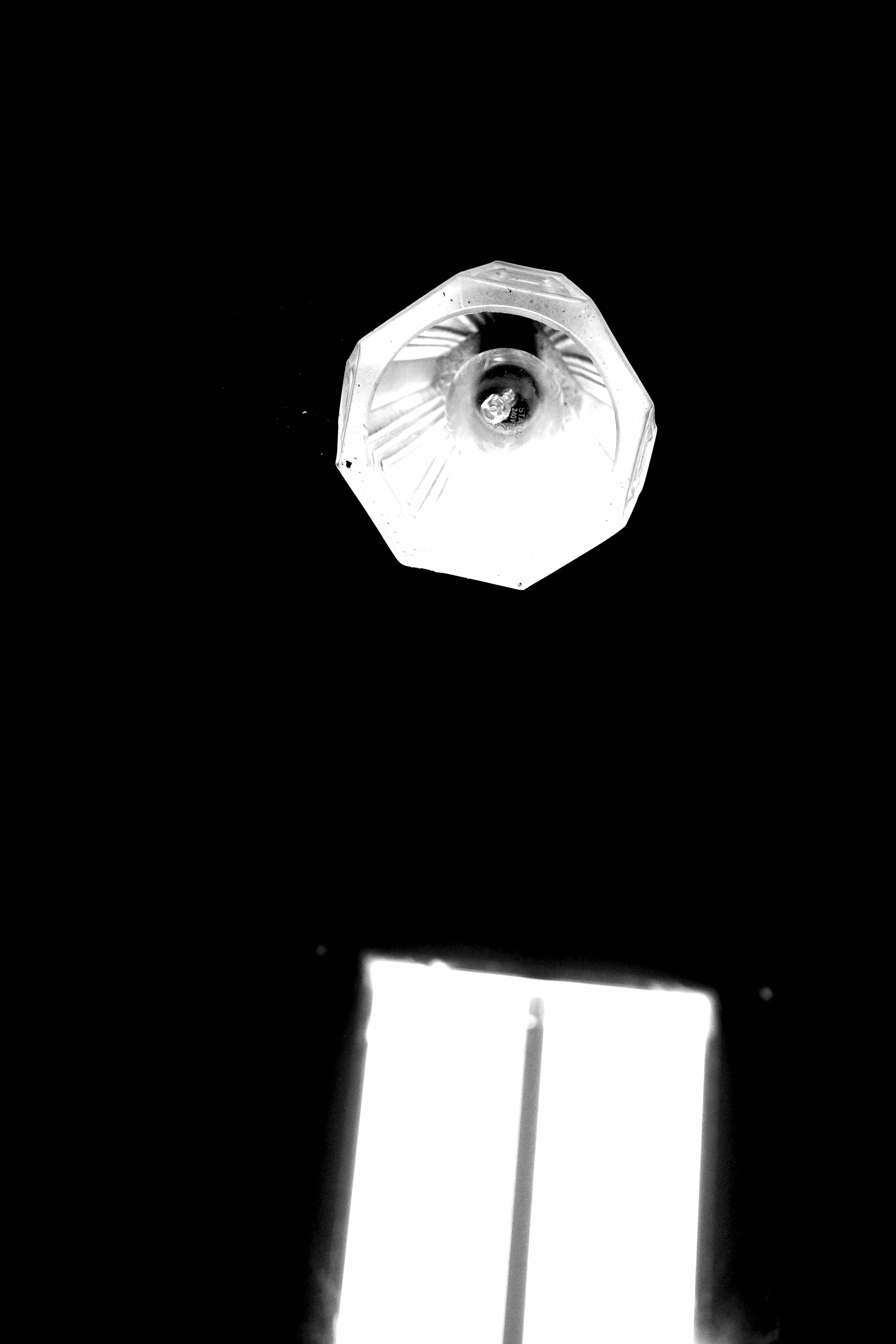 Black and white photograph of a window with a lampshade hanging in front of it. The lampshade has a geometric, faceted shape and is illuminated from within, creating a glow against the dark background.