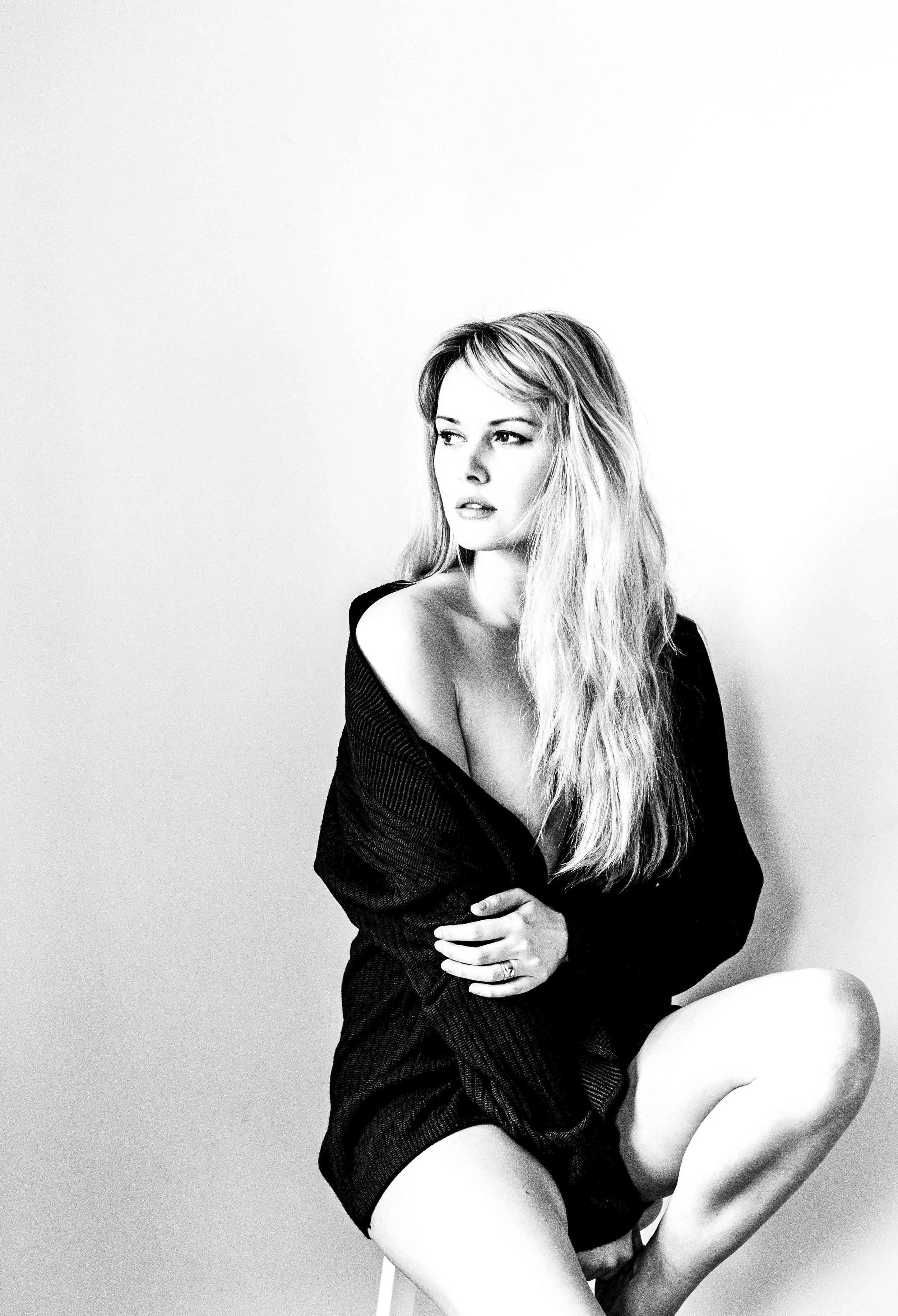 Black and white photo of a woman with long, wavy hair, sitting with one knee bent, wearing a loose, off-the-shoulder black top.