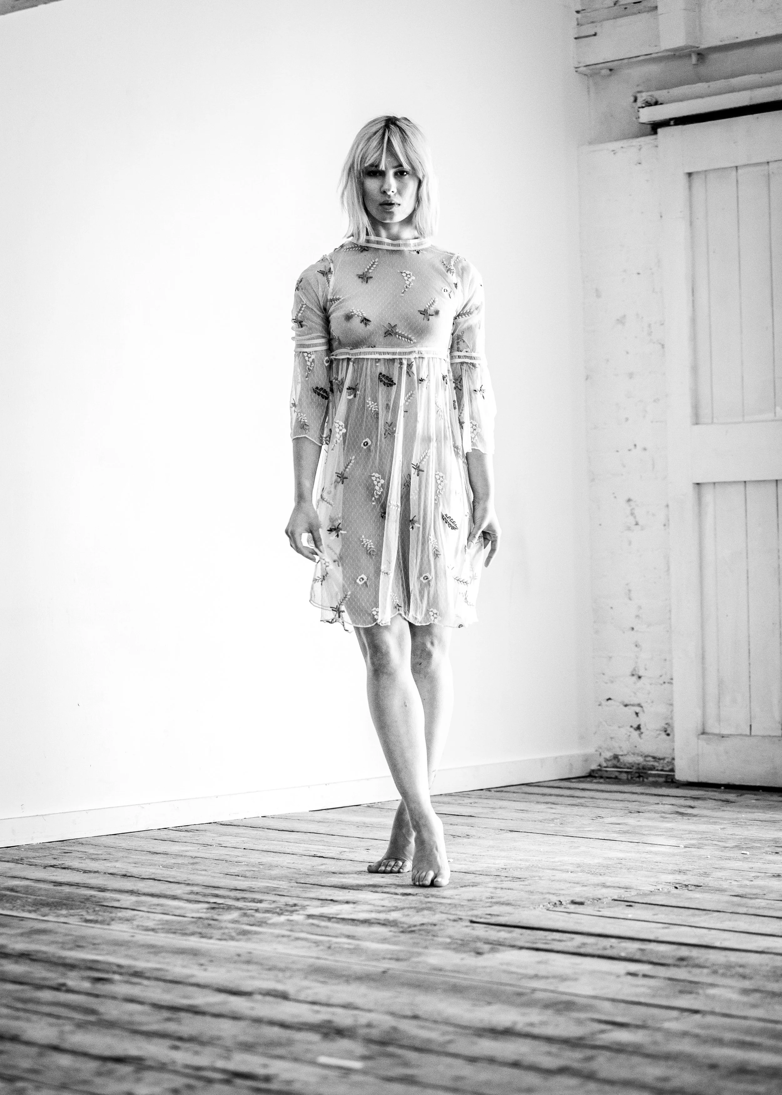 A woman with shoulder-length blonde hair standing barefoot on a wooden floor in a room with white walls. She is wearing a sheer, embroidered dress with long sleeves.