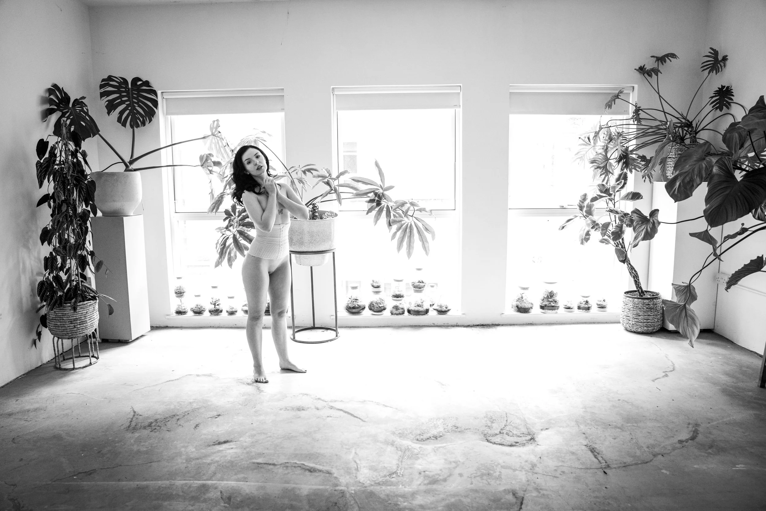 Black and white photo of a woman standing in a bright room with large windows, surrounded by potted plants on the windowsills and floor, with a bare concrete floor and minimal decor.