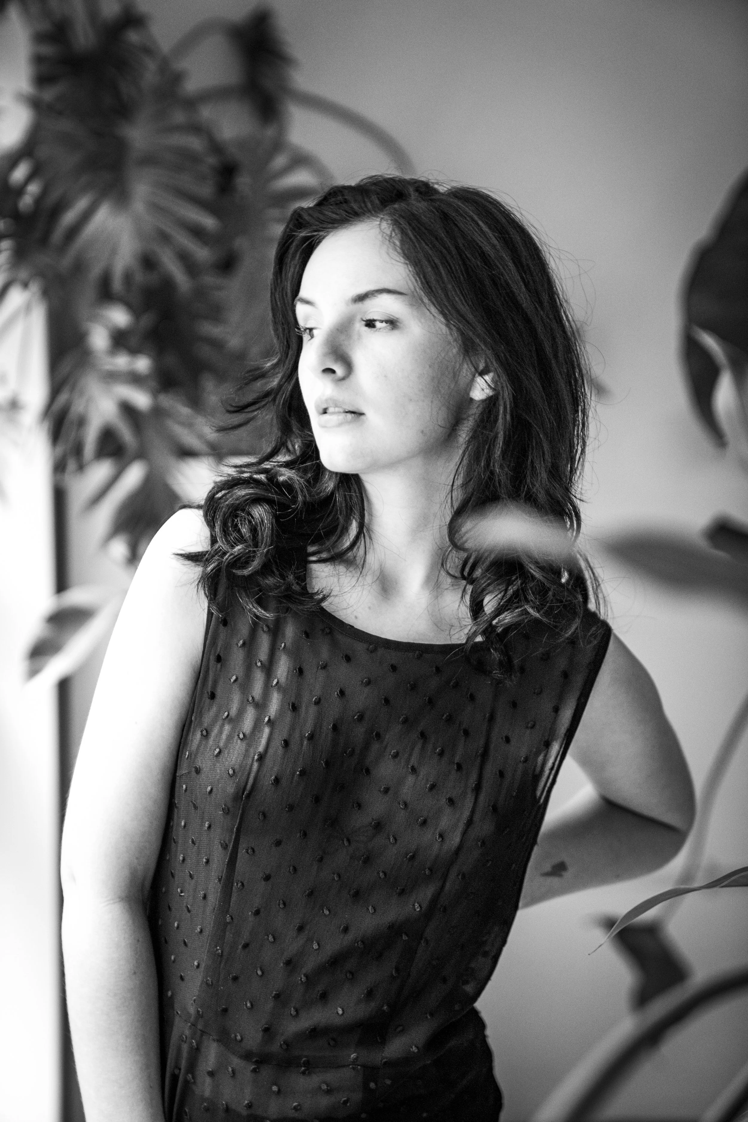 A woman with dark, wavy hair wearing a sleeveless, semi-transparent black top with small embellishments, standing near large plants, looking to her left, in black and white.