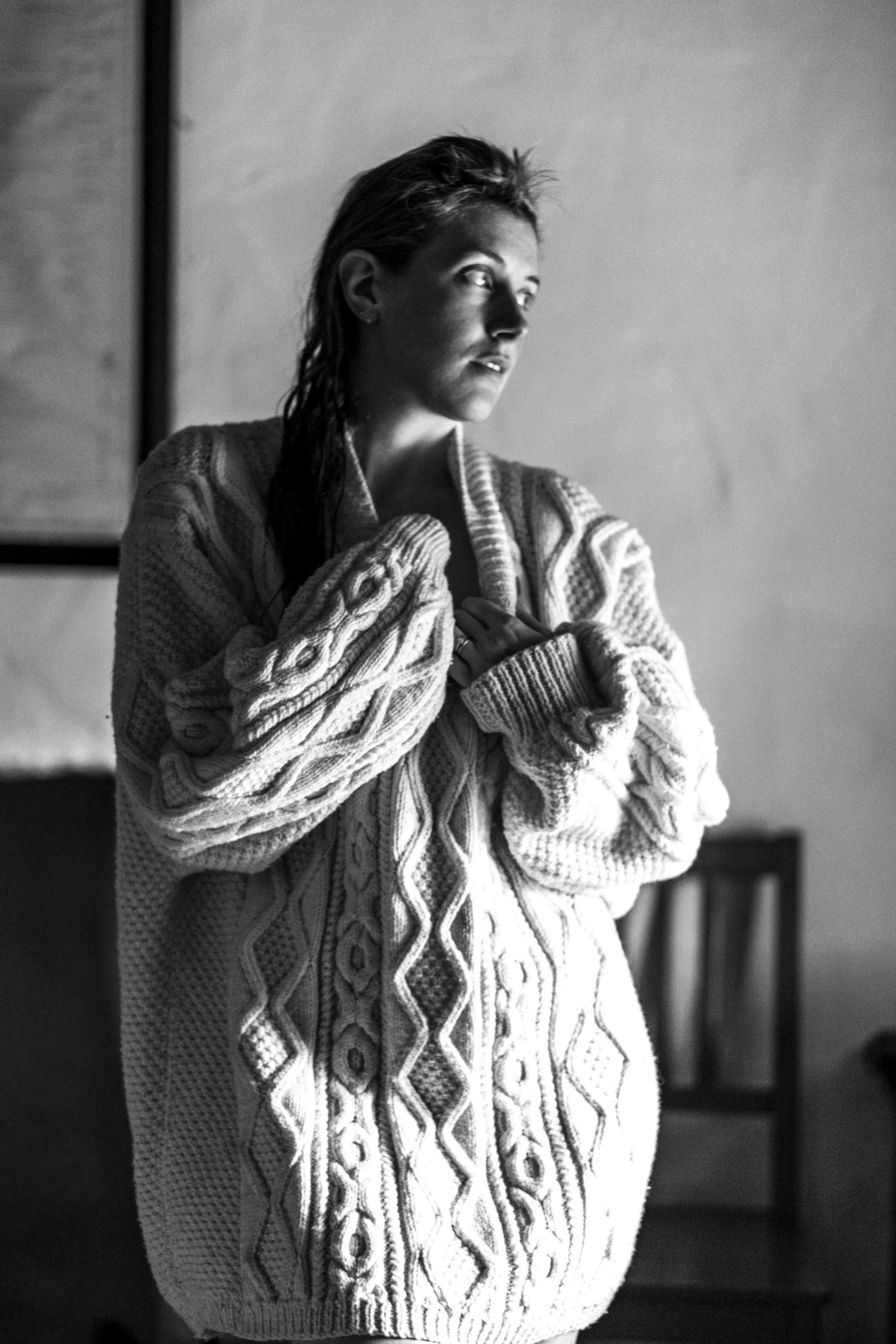 Black and white photo of a woman in a patterned sweater, gazing to her left with a contemplative expression, standing indoors near a wall.