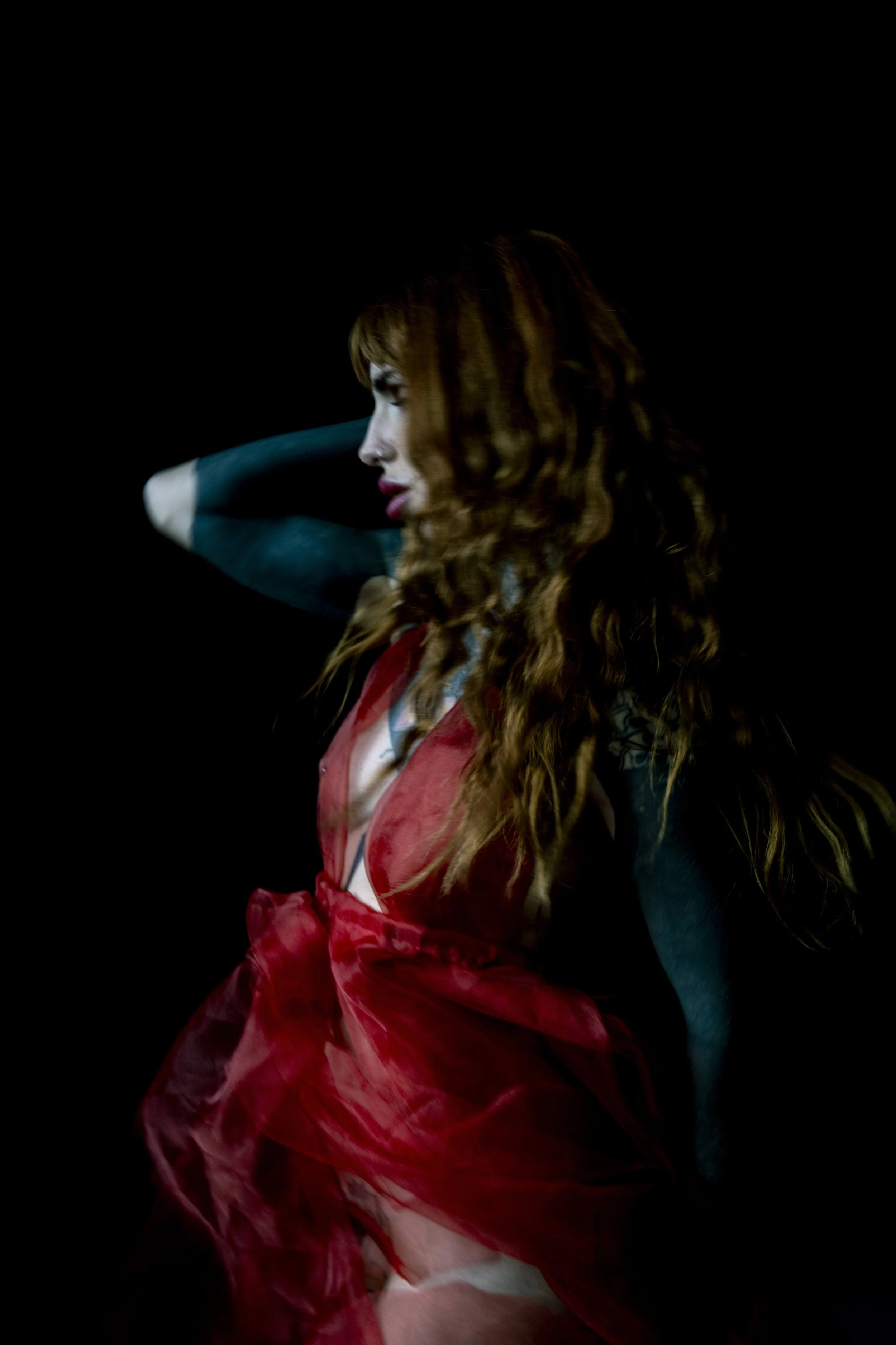 A young woman with long, curly red hair and fair skin is wearing makeup with lipstick. She is dressed in a red, semi-transparent, flowing garment and is posed with her eyes closed and one arm raised behind her head, against a black background.