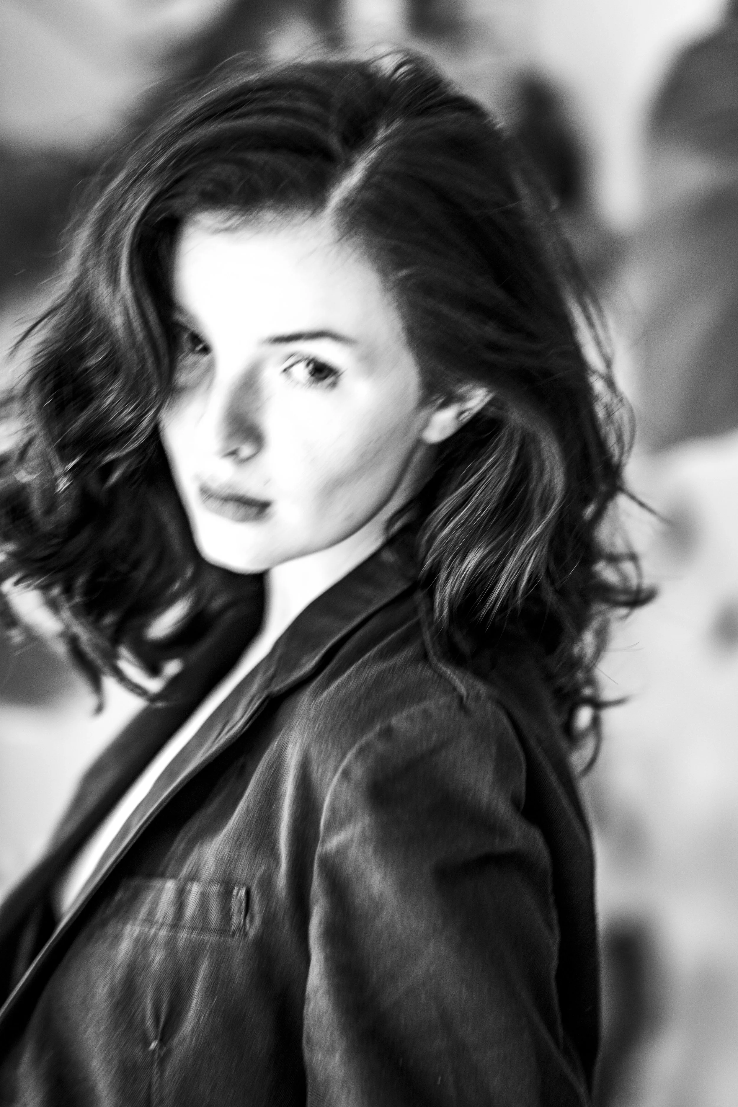 A black and white close-up portrait of a woman with wavy hair, wearing a denim jacket, looking over her shoulder.