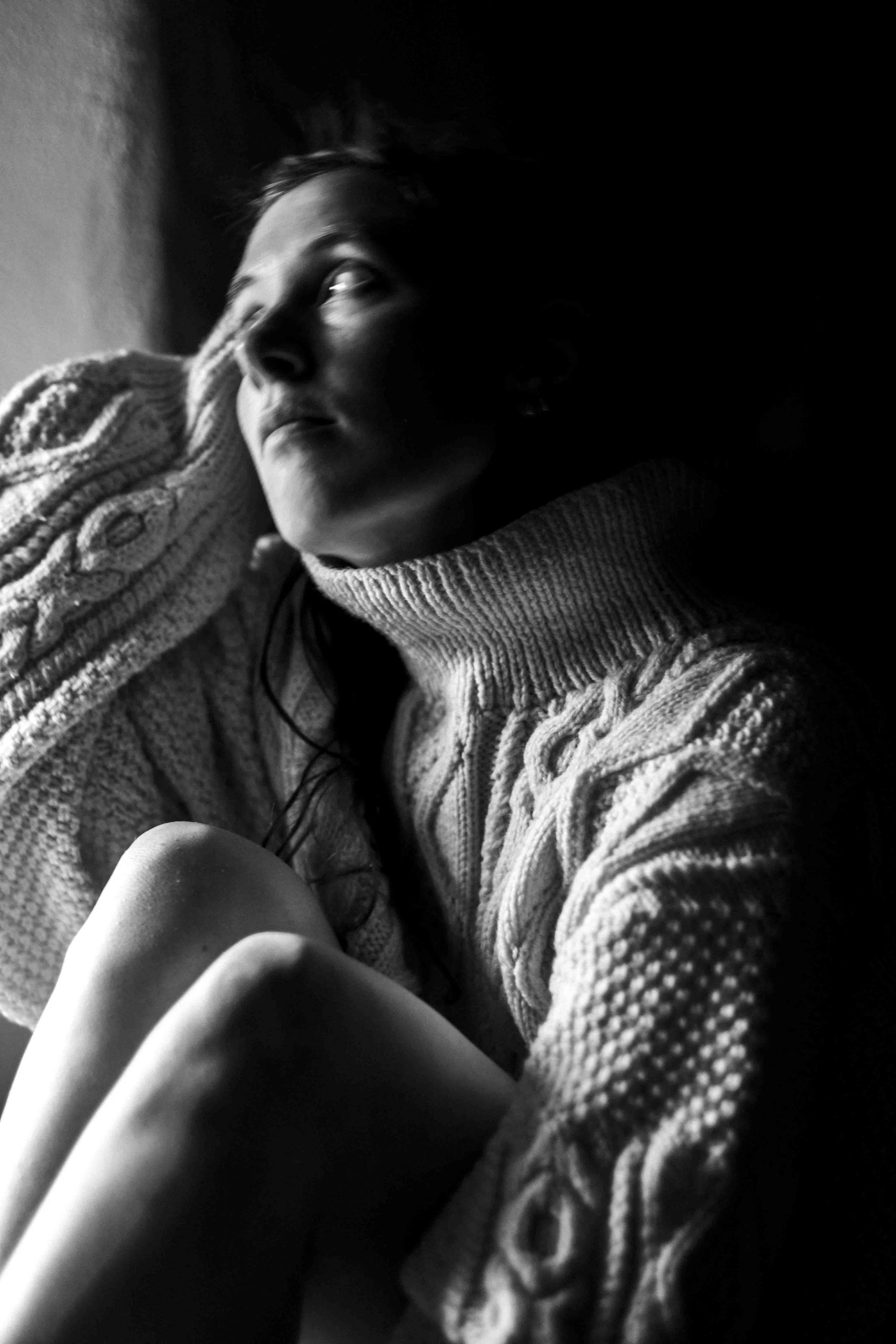 A black and white photo of a woman lying on her side, looking up, with her head resting on her hand, wearing a cable-knit sweater.