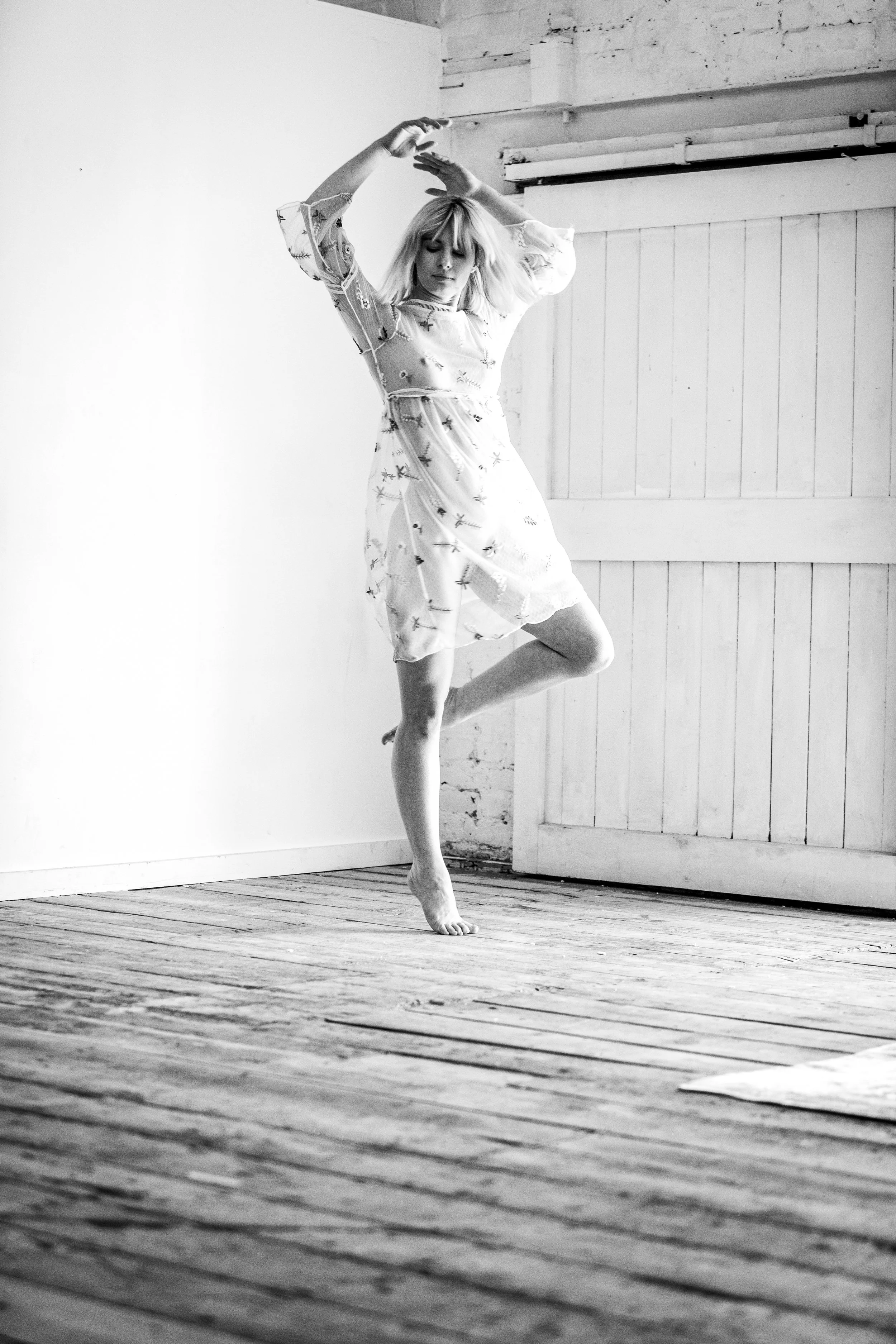 A woman dancing on a wooden floor in a minimally decorated room with white walls and a wooden door.