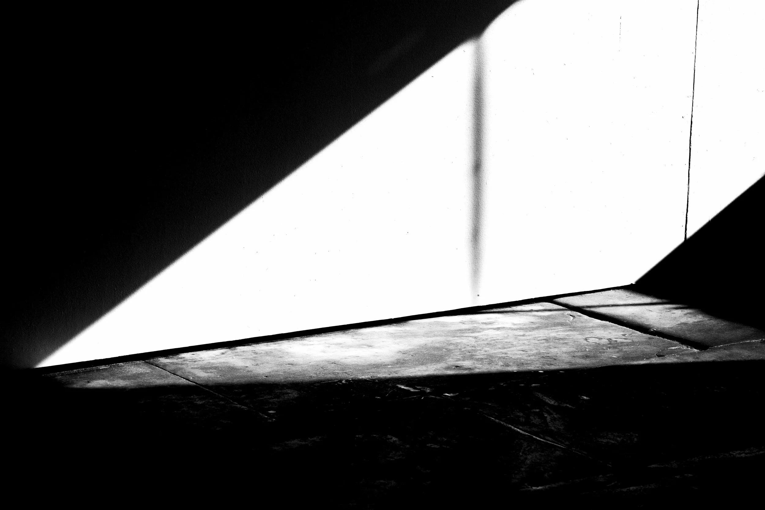 A black and white photo of a shadowed wall with light coming through a window, casting shadows of window bars on the wall and floor.
