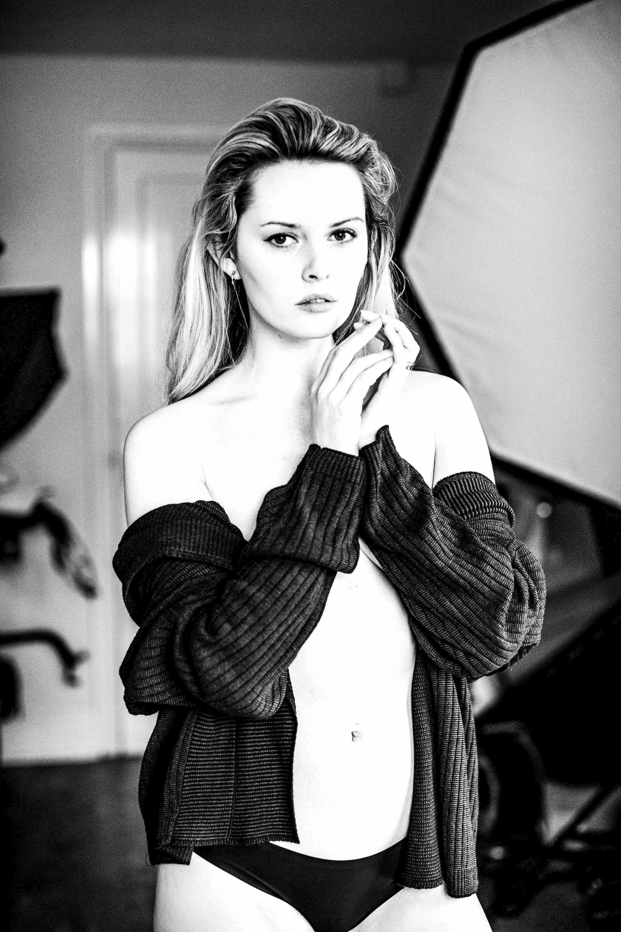 Black and white photo of a woman with long, wavy hair and striking features, wearing a partially open plaid shirt off her shoulders, revealing her bare midriff and underwear, in an indoor setting with a blurred background.