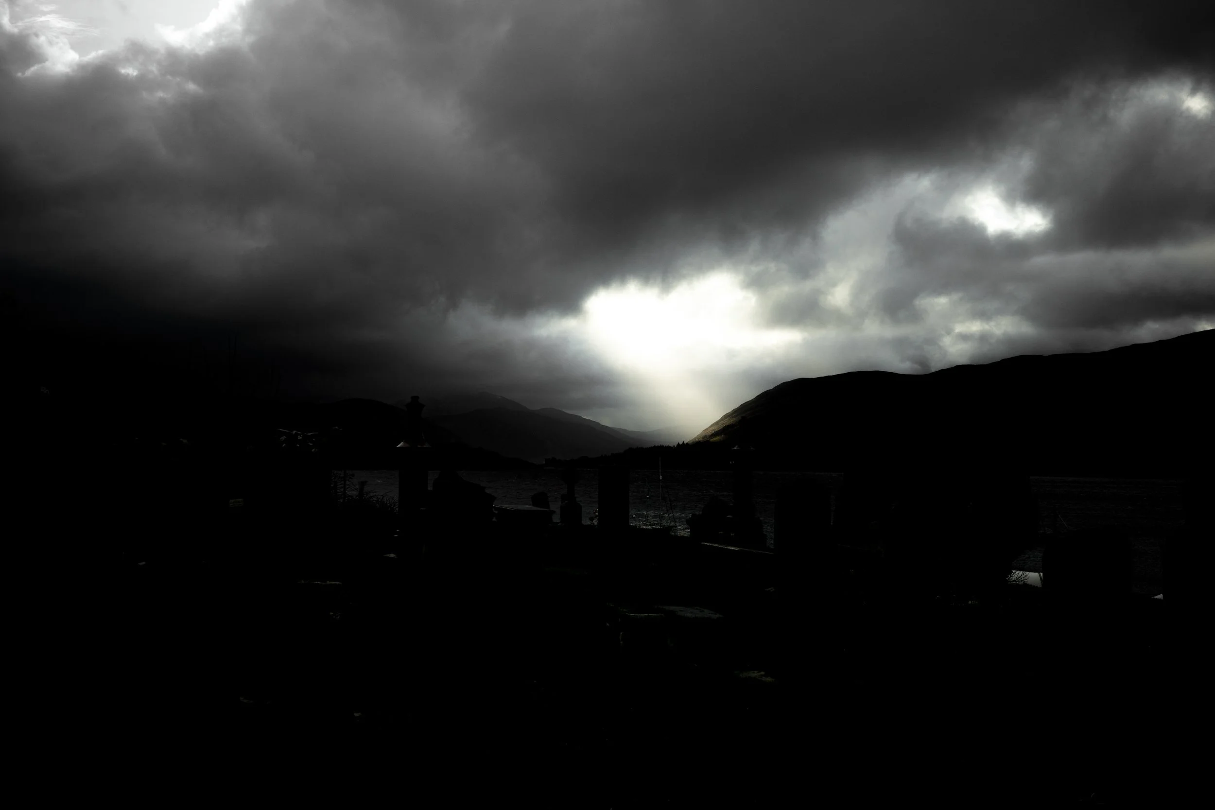 Dark storm clouds over mountainous landscape with faint sunlight breaking through.
