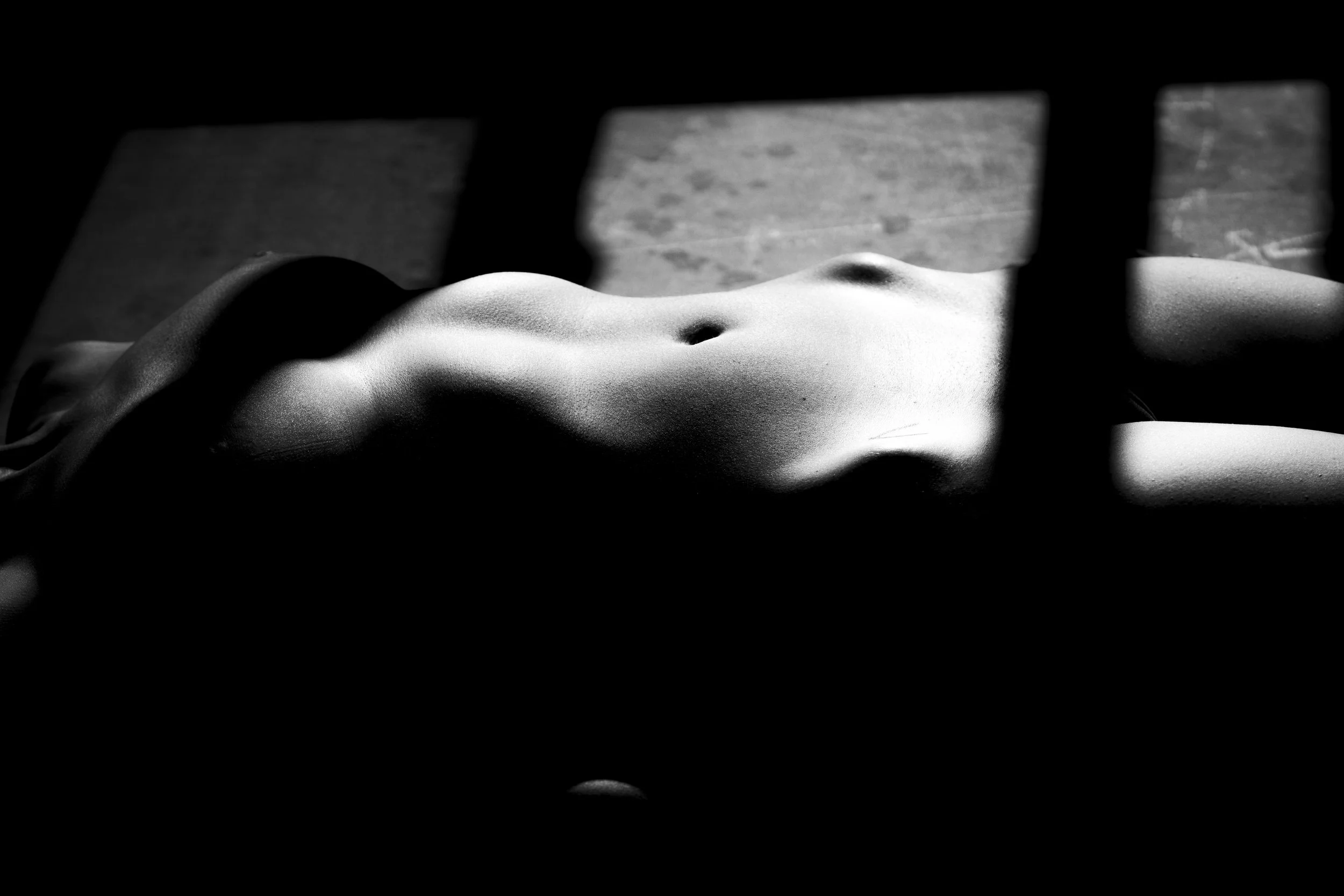 A black and white photograph of a human torso lying on a surface, partially obscured by shadows and horizontal bars.