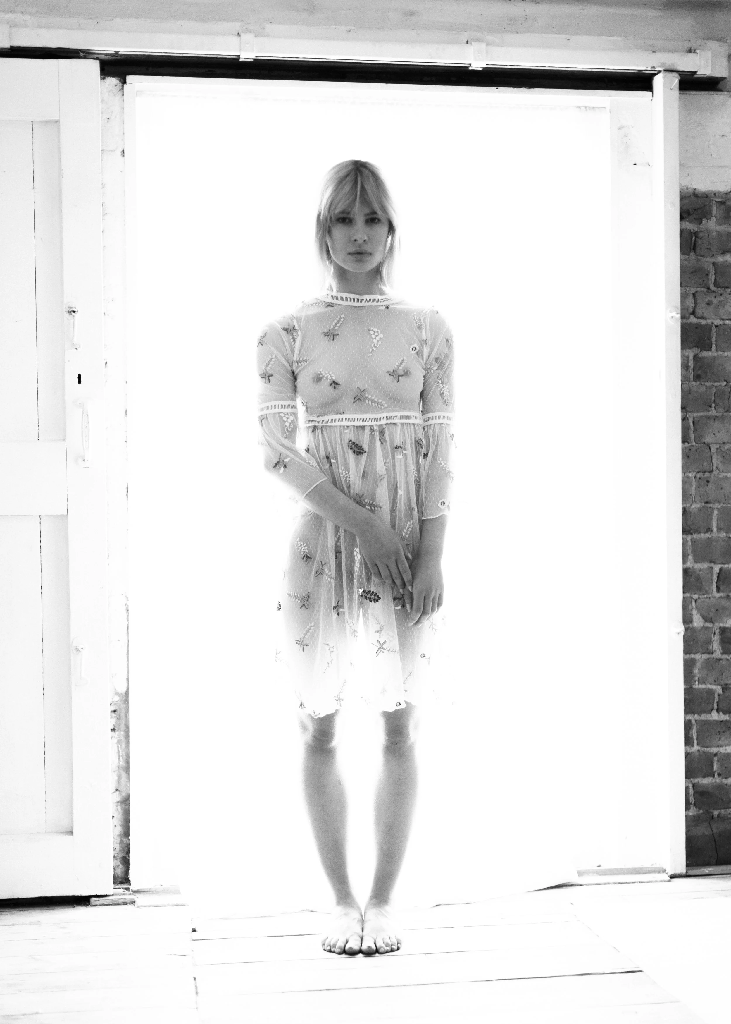A woman standing upright with her hands down by her sides, facing forward, in front of a bright, backlit background, wearing a patterned dress with embroidery, with bare feet on a wooden floor, next to a brick wall.