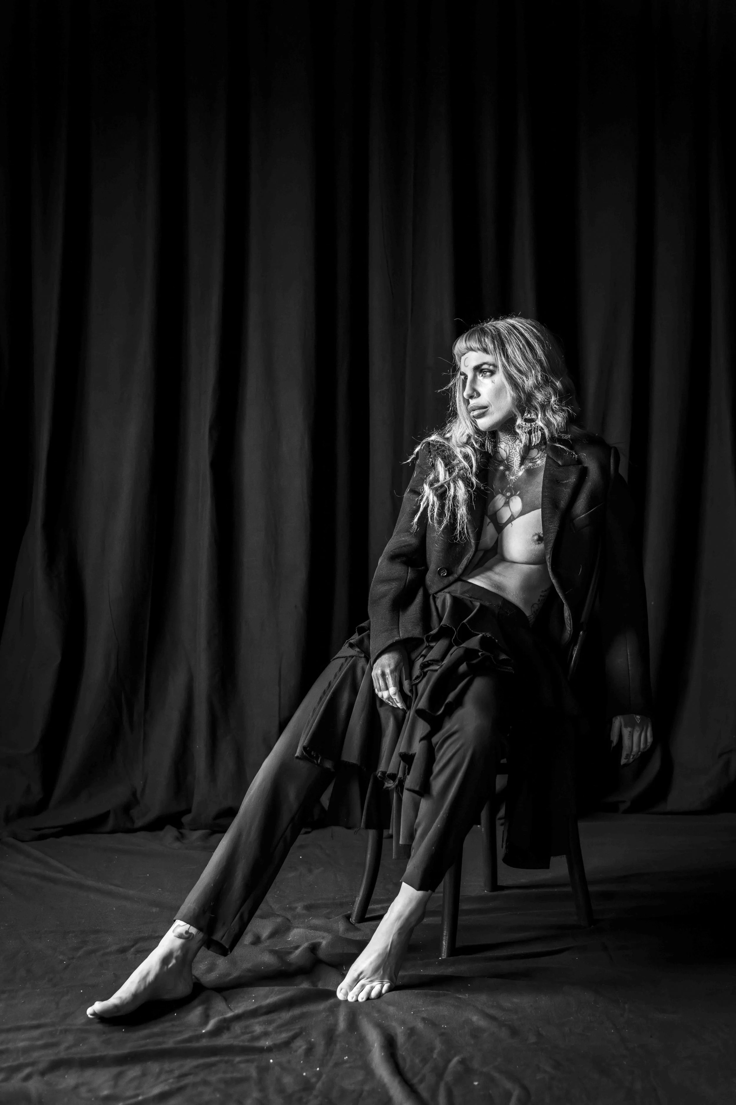 A woman with wavy hair and tattoos seated on a chair in front of a dark curtain, wearing a blazer and pants, with her hand resting on her knee and legs crossed, in a black and white photo.