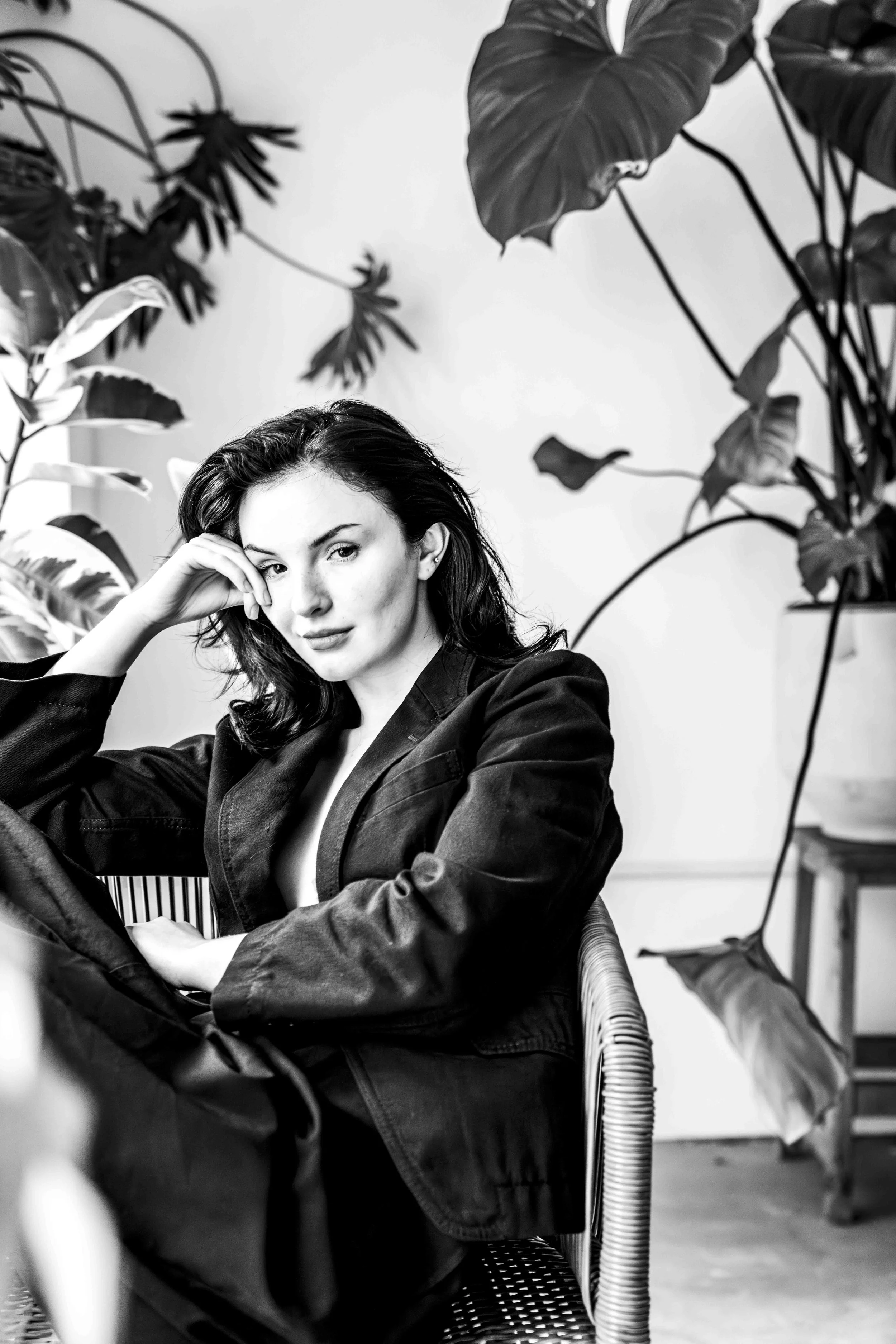 Black and white photo of a woman with dark hair sitting on a wicker chair, surrounded by large tropical plants, looking at the camera with a relaxed pose and a hand resting on her face.