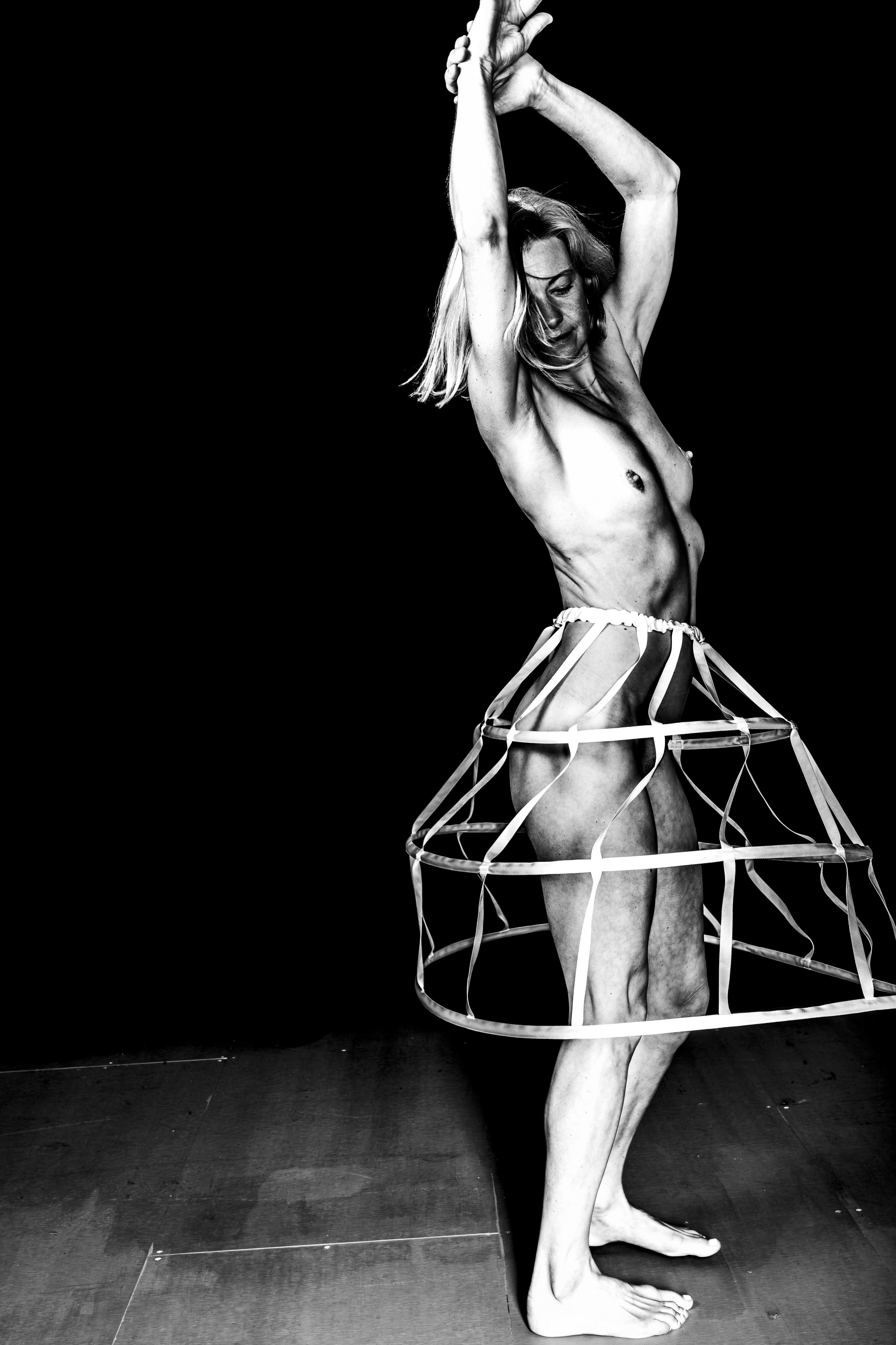 Black and white photo of a woman standing with her arms raised above her head, wearing a lightweight, open wireframe skirt, topless, on a dark background.