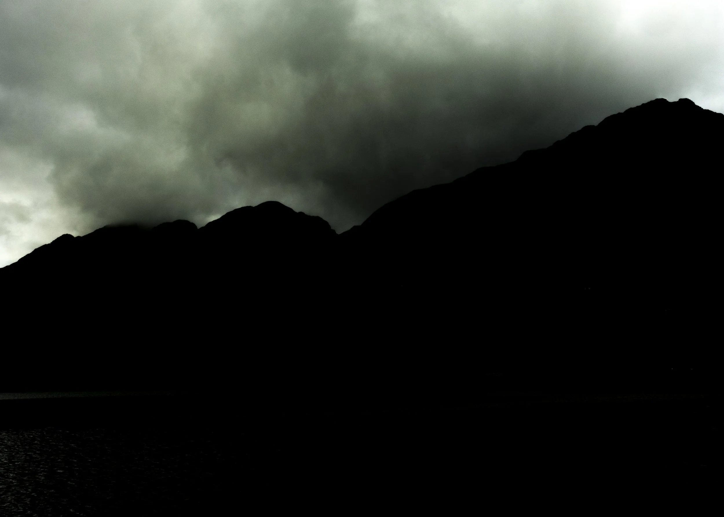 Dark, cloudy sky over a mountain silhouette with a body of water in the foreground.