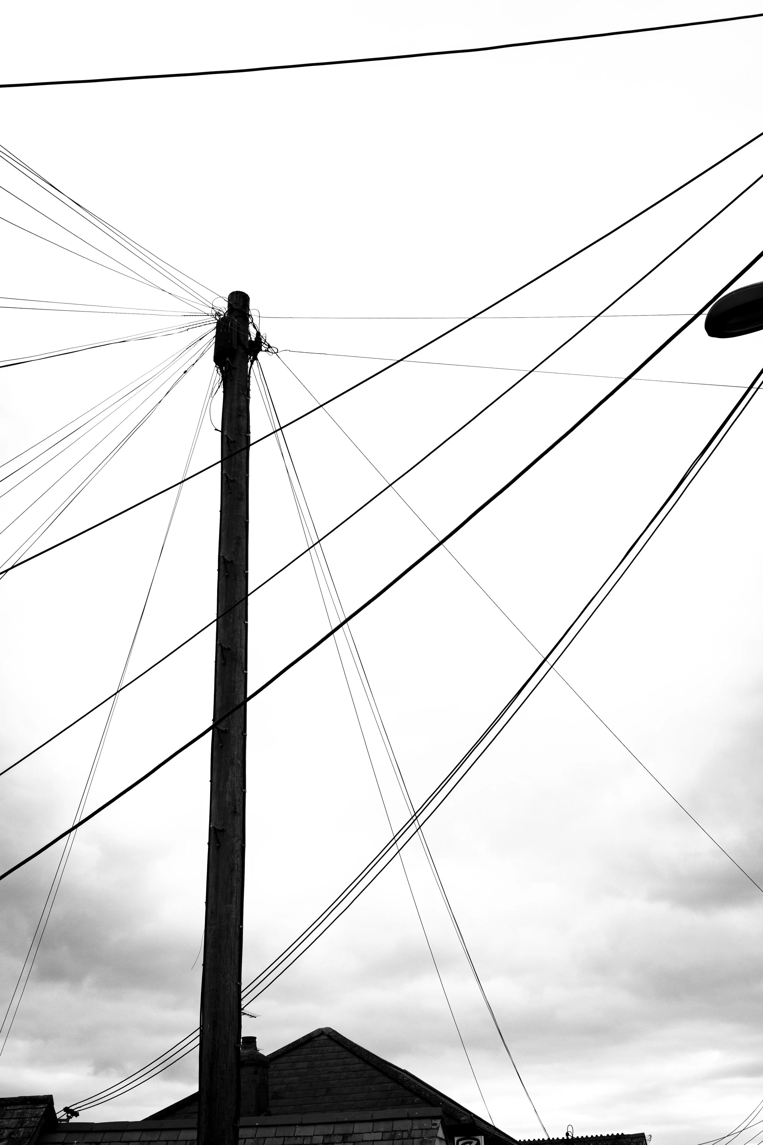 A tall utility pole with wires extending in various directions against a cloudy sky, with rooftops visible at the bottom of the image.