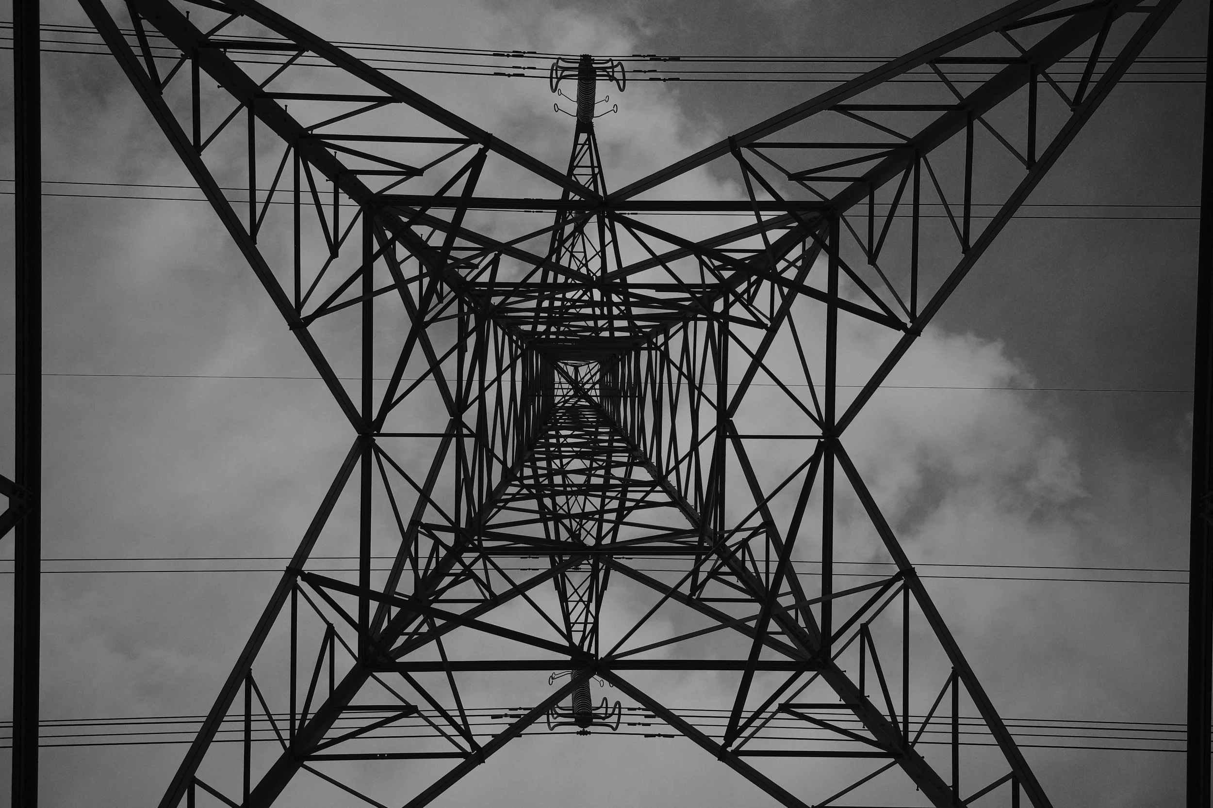 Looking up at an electrical transmission tower from below, showing the metal framework and power lines against a cloudy sky.