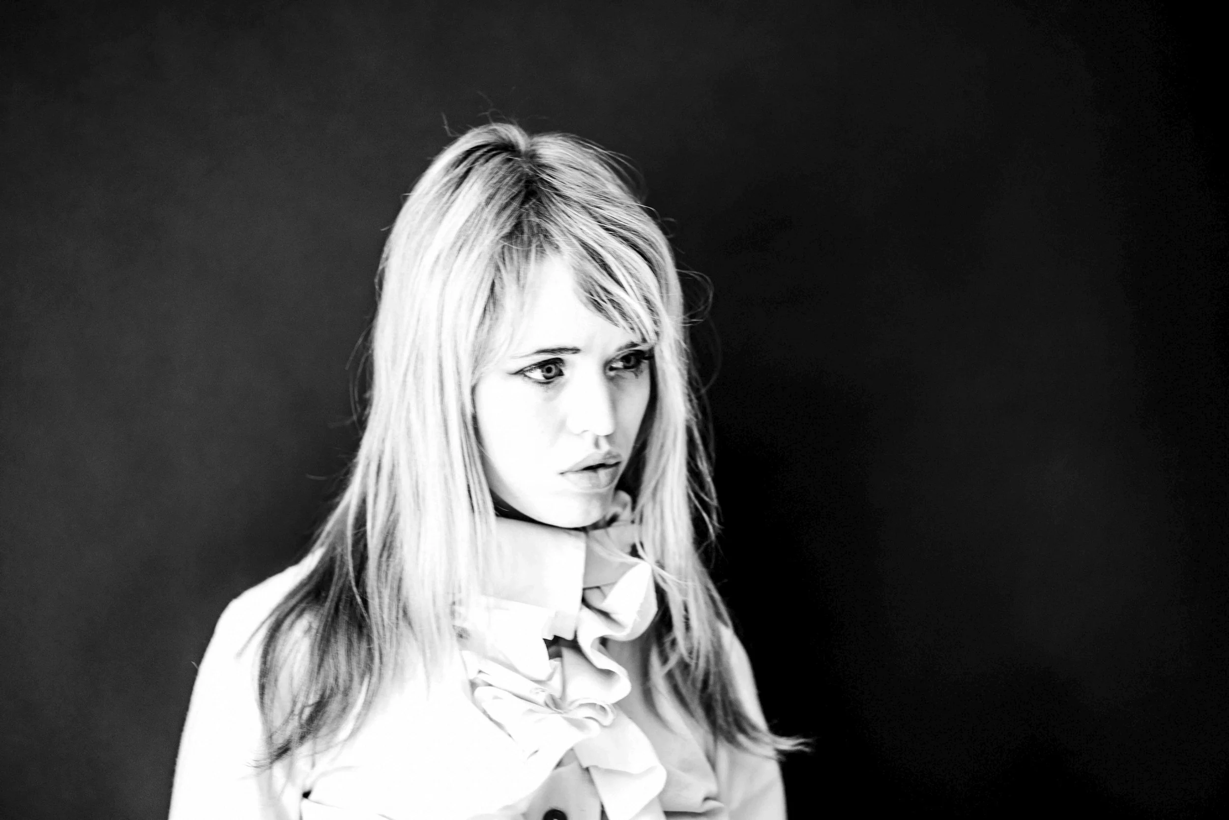 Black and white photo of a woman with long hair, wearing a collared shirt and a bow tie, looking slightly to her left against a dark background.