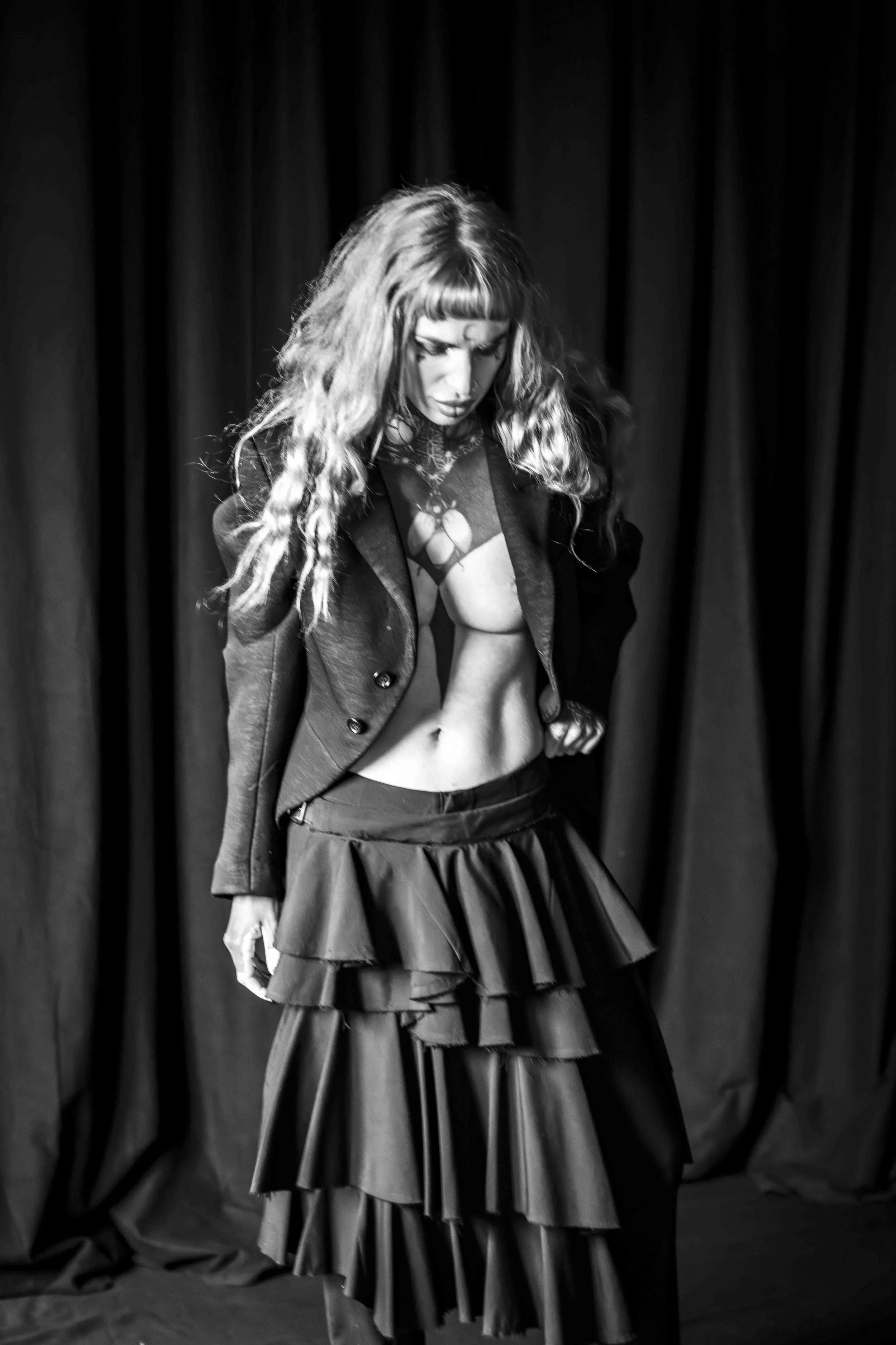 Black and white photo of a woman with long curly hair, wearing a blazer and a layered skirt, standing in front of a dark curtain, looking down.