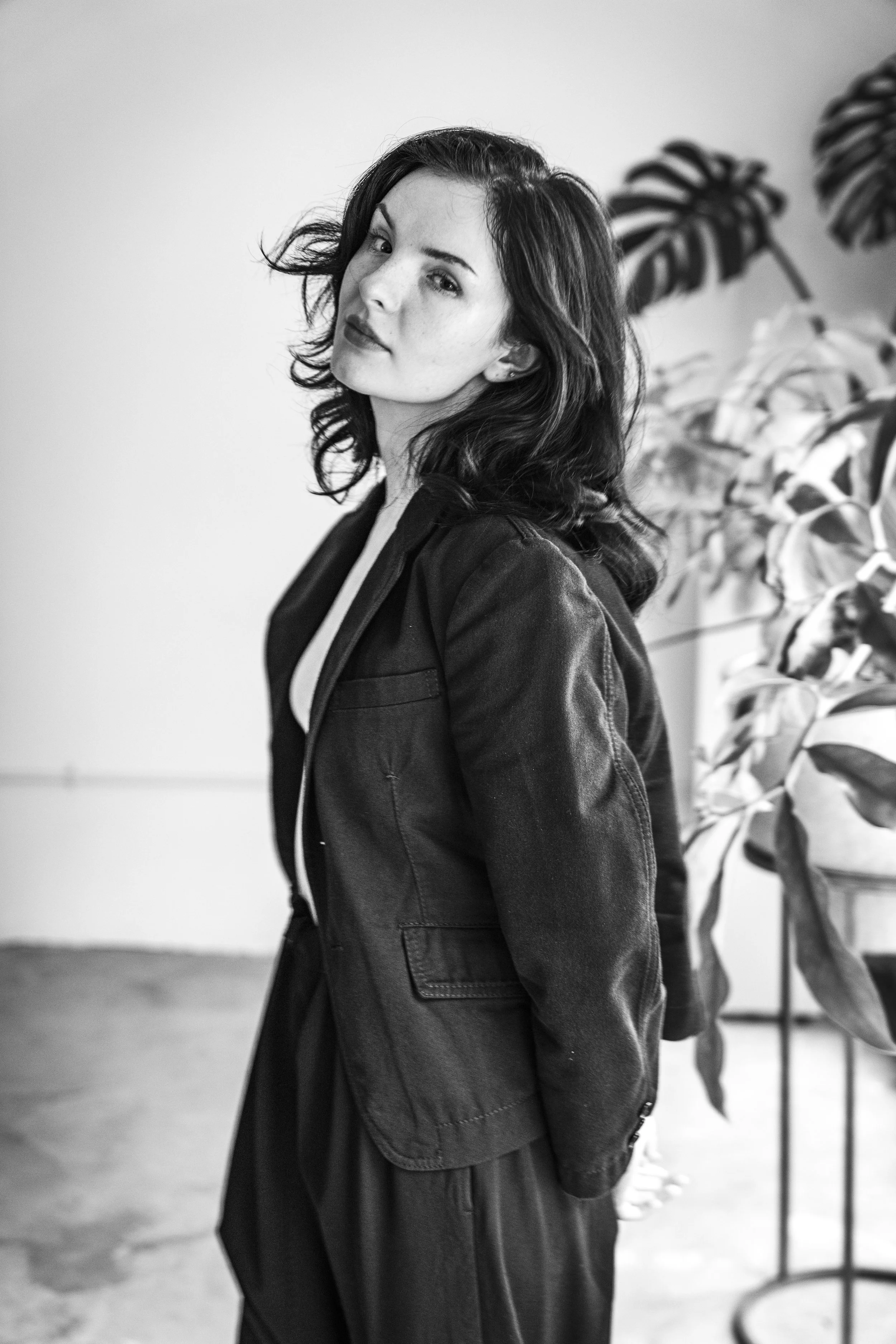 Black and white portrait of a young woman with wavy dark hair, wearing a dark jacket, standing in front of a plain wall with a large potted plant to her right.