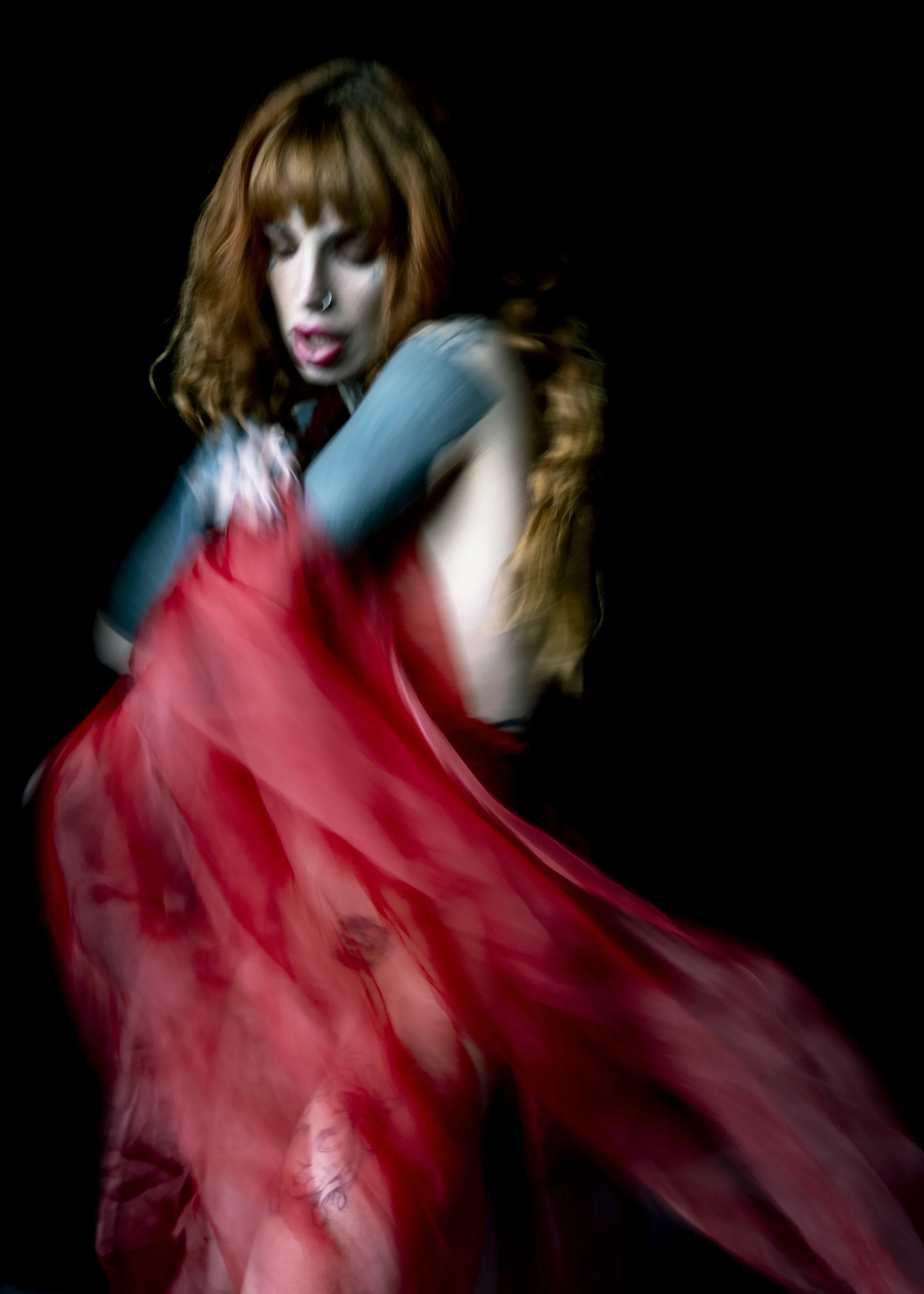 A woman with red hair and makeup features, wearing a red dress, is captured in motion against a black background.