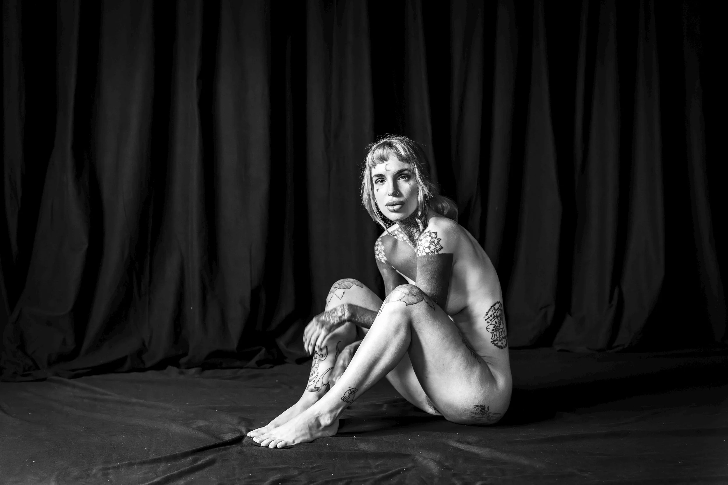 A person with numerous tattoos sitting on a stage with black curtains in the background, lit from above, in black and white.