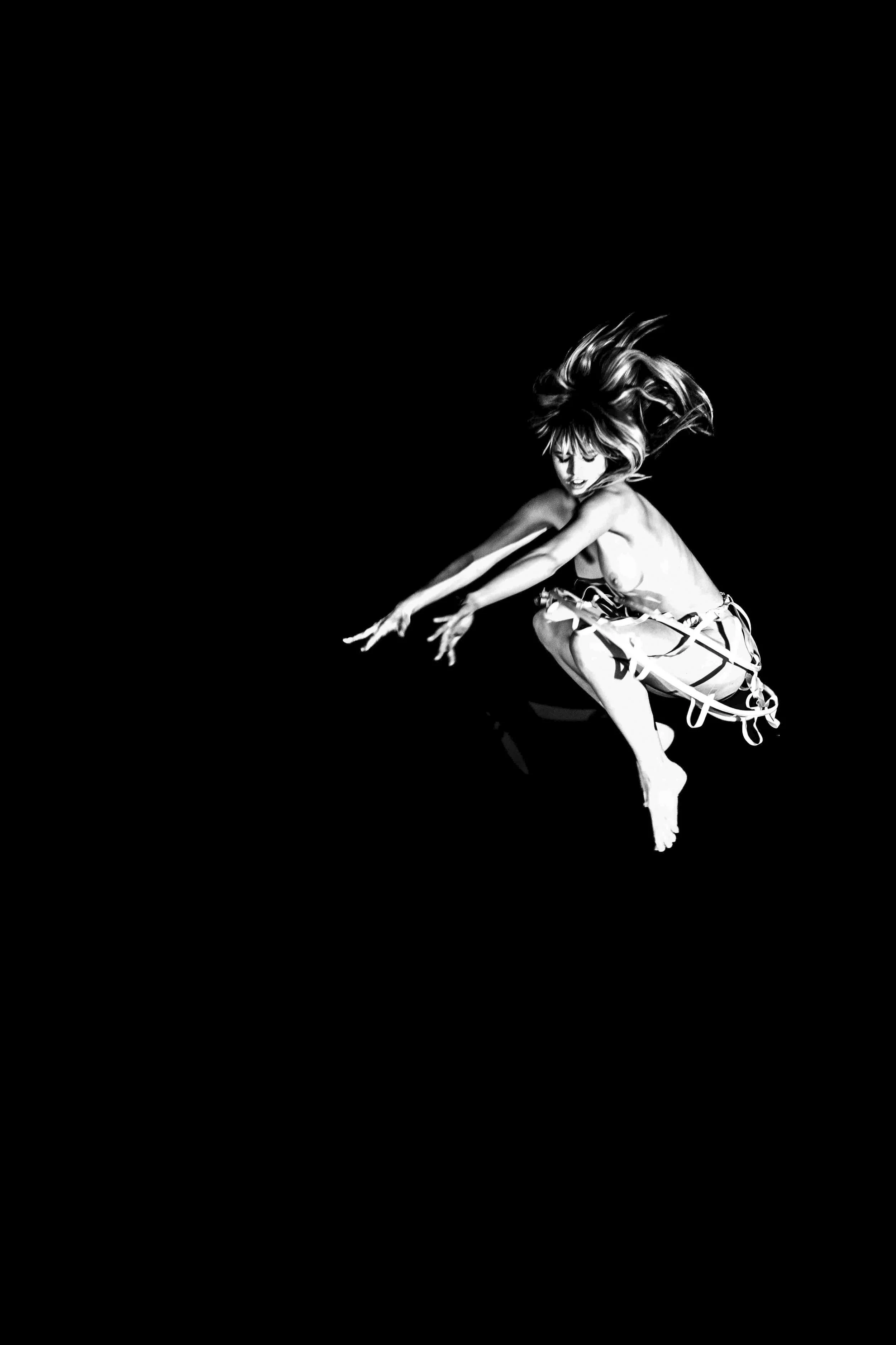 Black and white image of a woman with hair flowing, squatting with arms extended forward, wearing a skirt with laces.