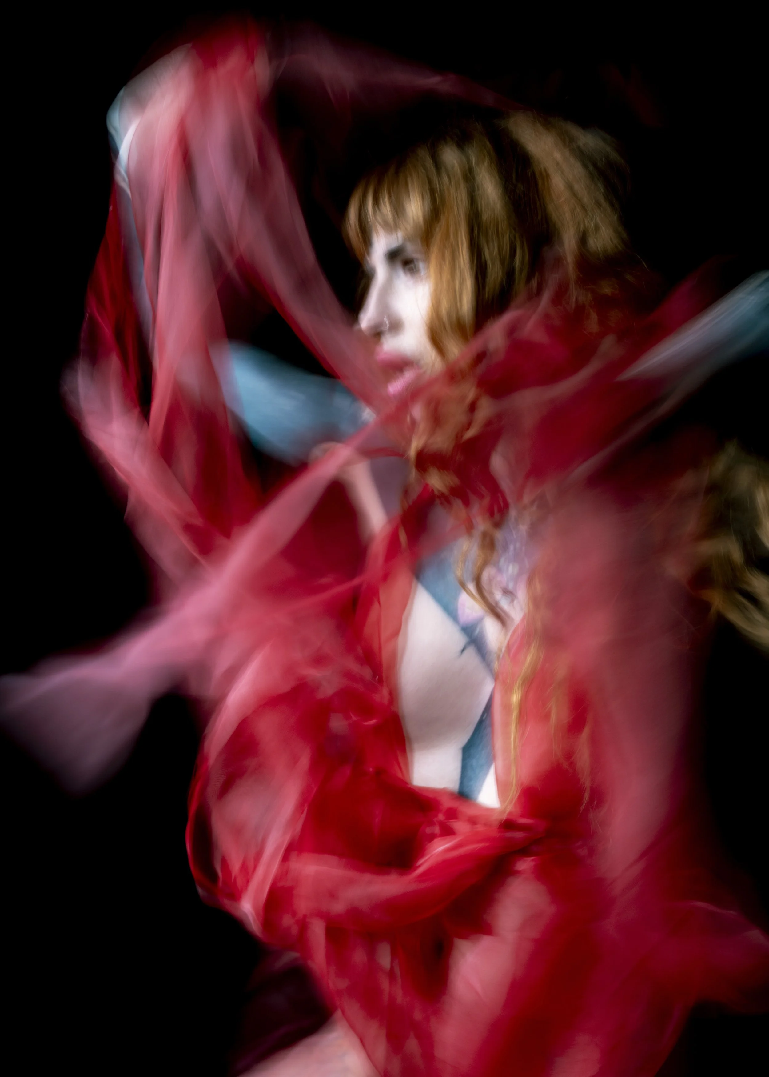 A woman with red hair is surrounded by flowing red fabric, creating a dynamic and artistic effect against a black background.