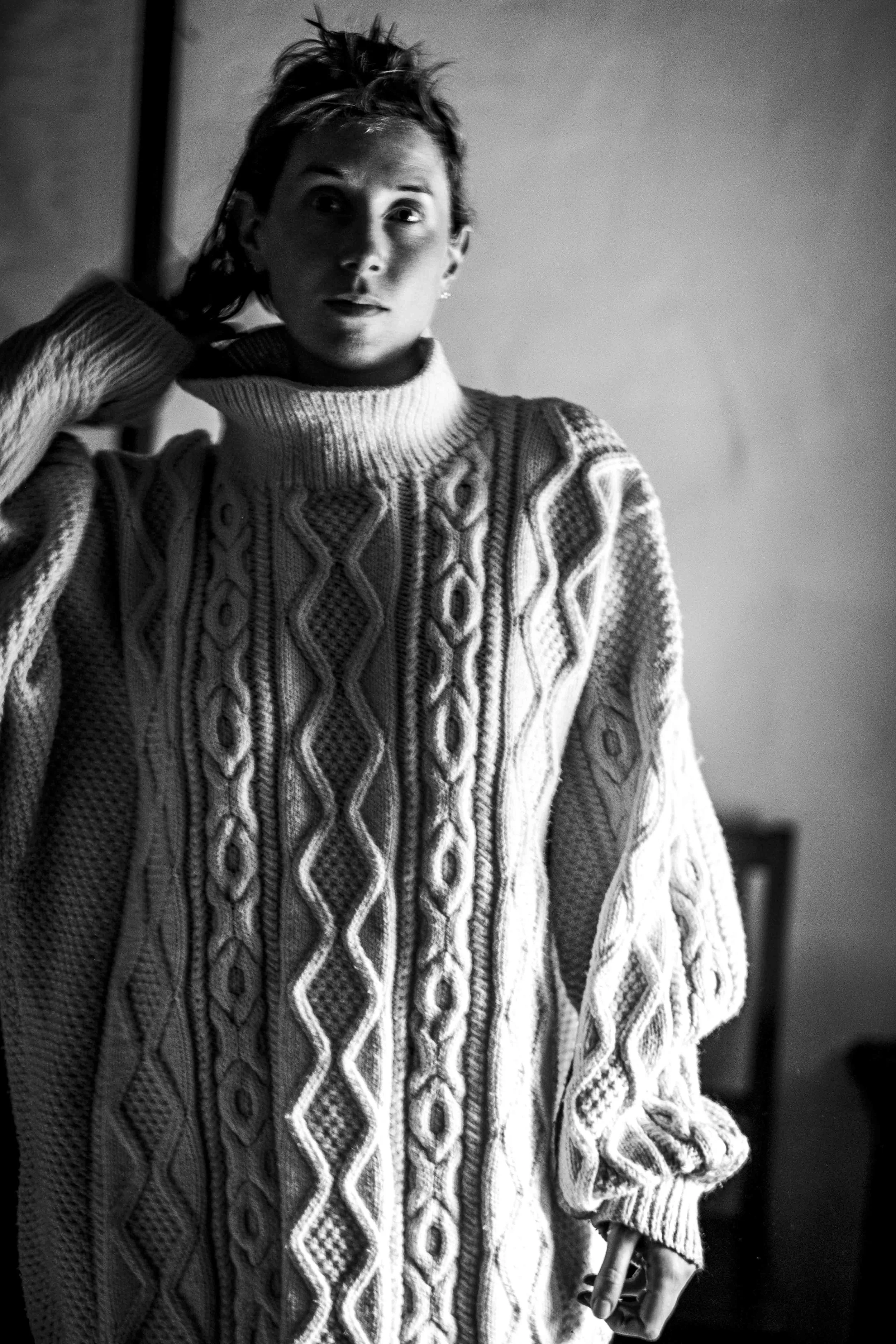 A woman with short hair wearing a cable-knit sweater, looking directly at the camera, holding a cigarette in her right hand.