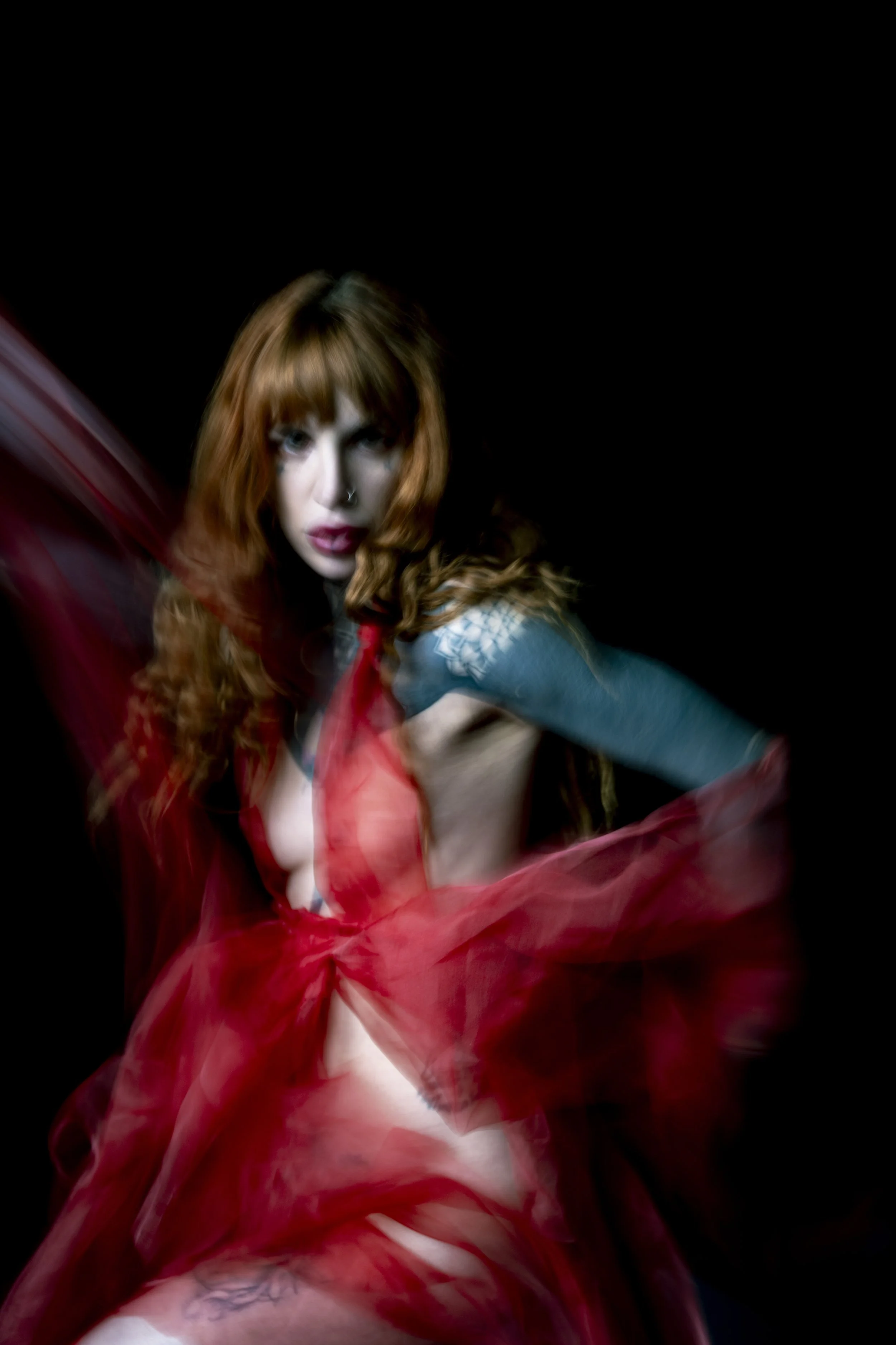 A woman with long red hair, wearing a red dress and a blue sleeve, poses against a black background with blurred red fabric swirling around her.