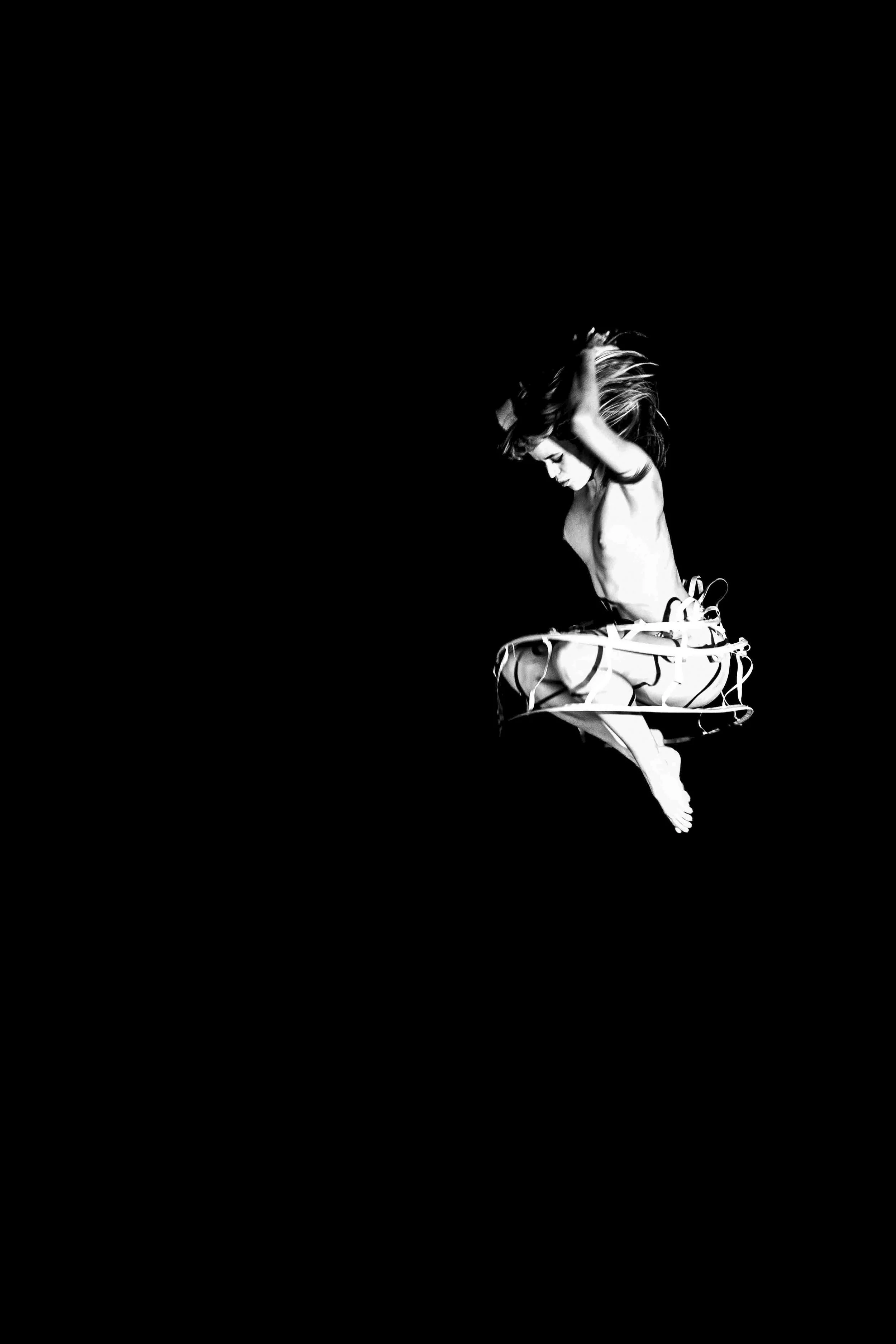 Black and white photo of a woman in a dynamic pose, with her hair flying and her arms raised above her head, against a dark background.