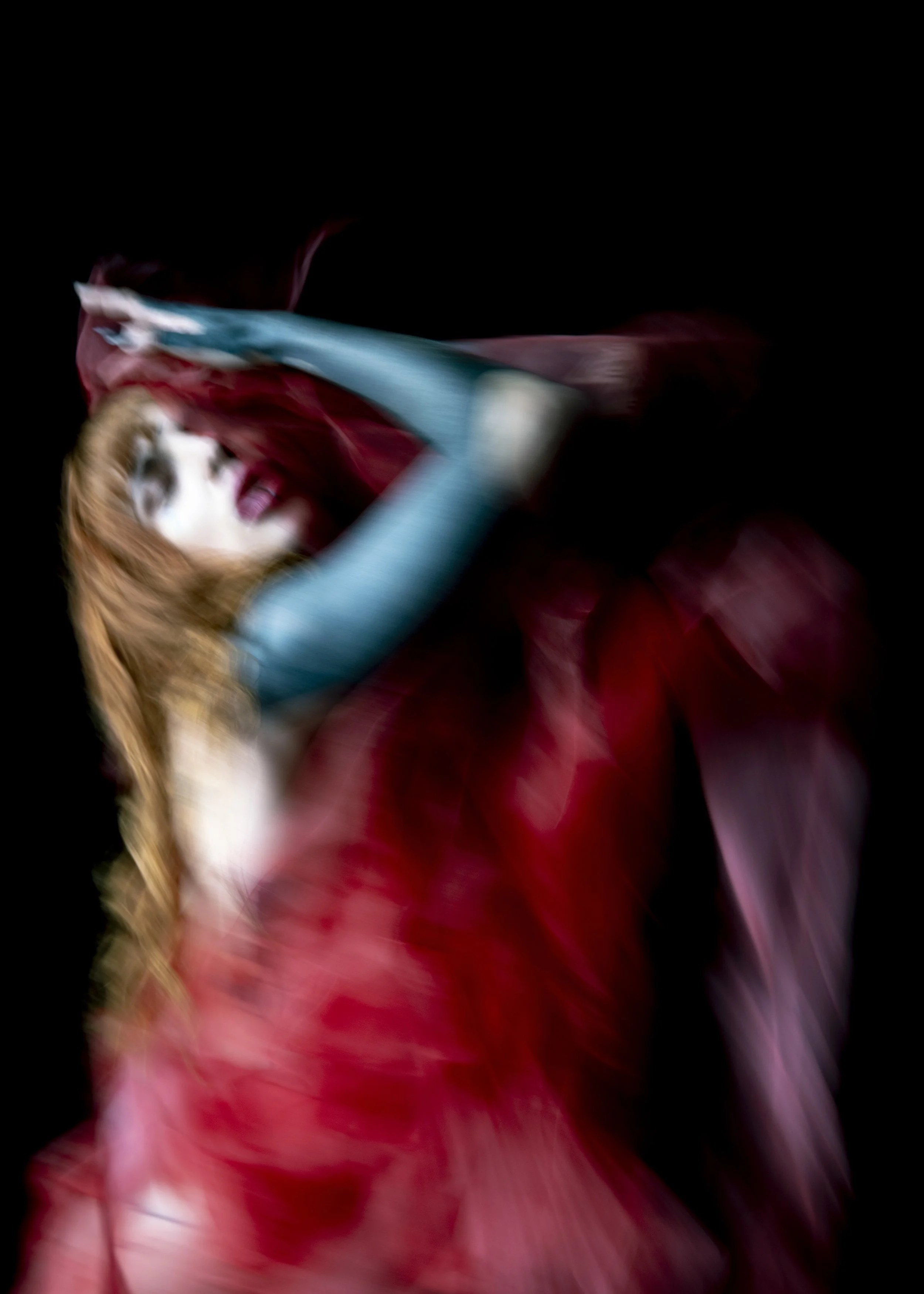 Blurred image of a woman with long hair wearing a red dress and blue gloves, with a dark background.