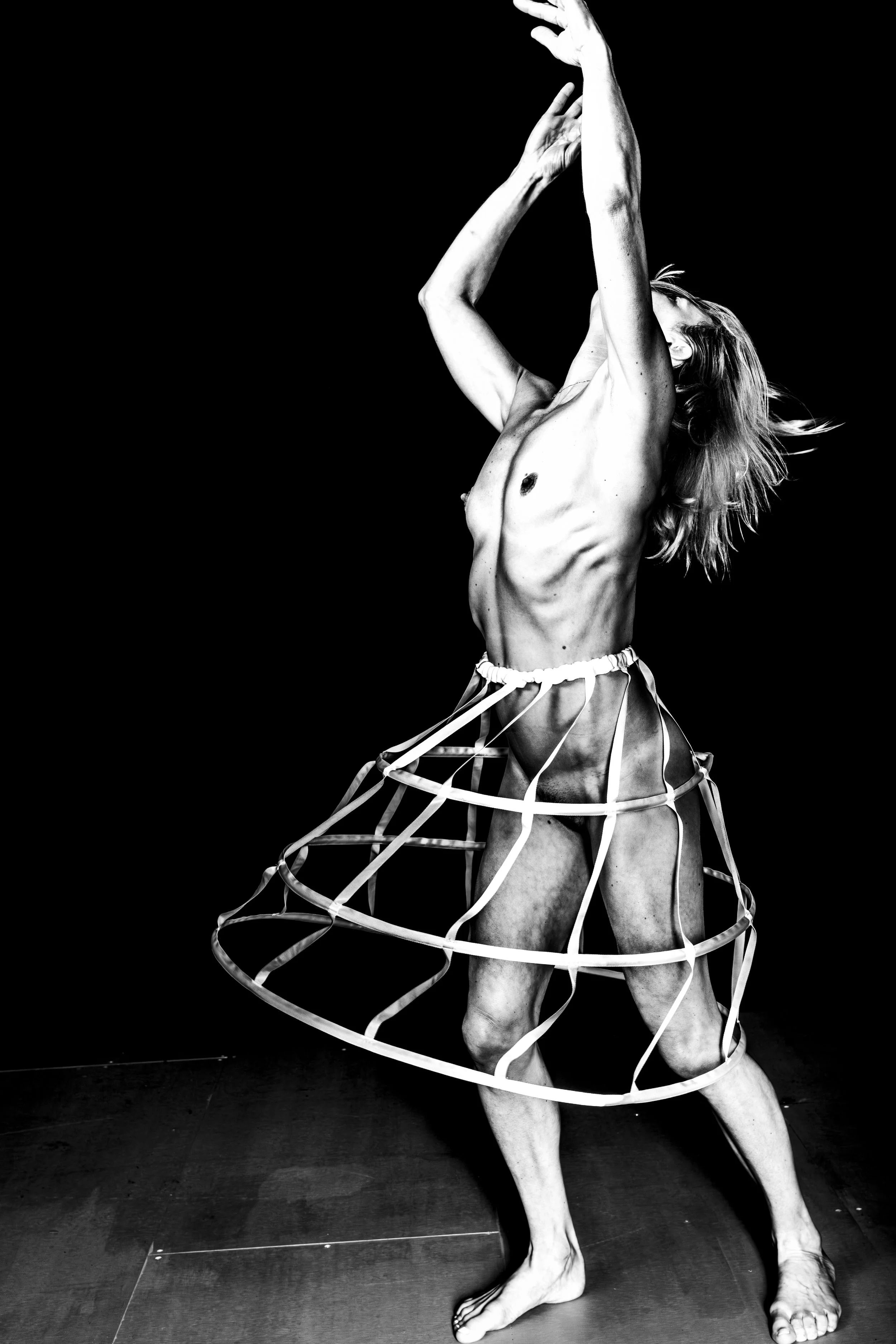 A black and white photo of a woman with long hair, topless, wearing a wide, geometric skirt, striking a dance pose with her arms raised and head tilted back.