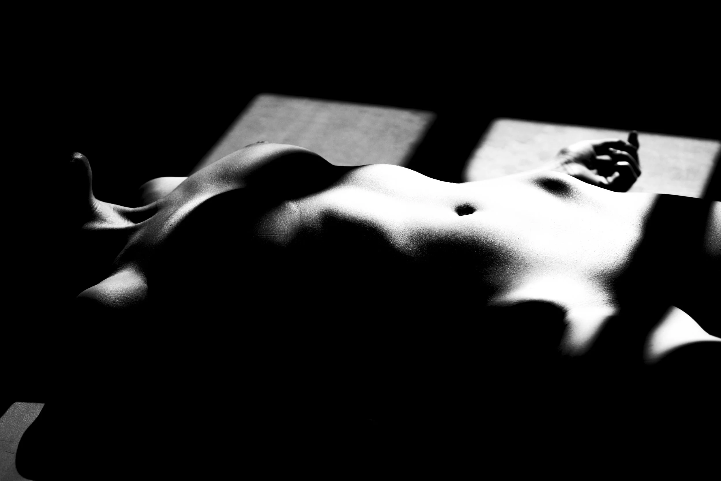 A black and white photograph of a reclining nude figure, lying on their back with one arm resting above their head, cast in shadow with sunlight creating dramatic highlights and shadows on the body.