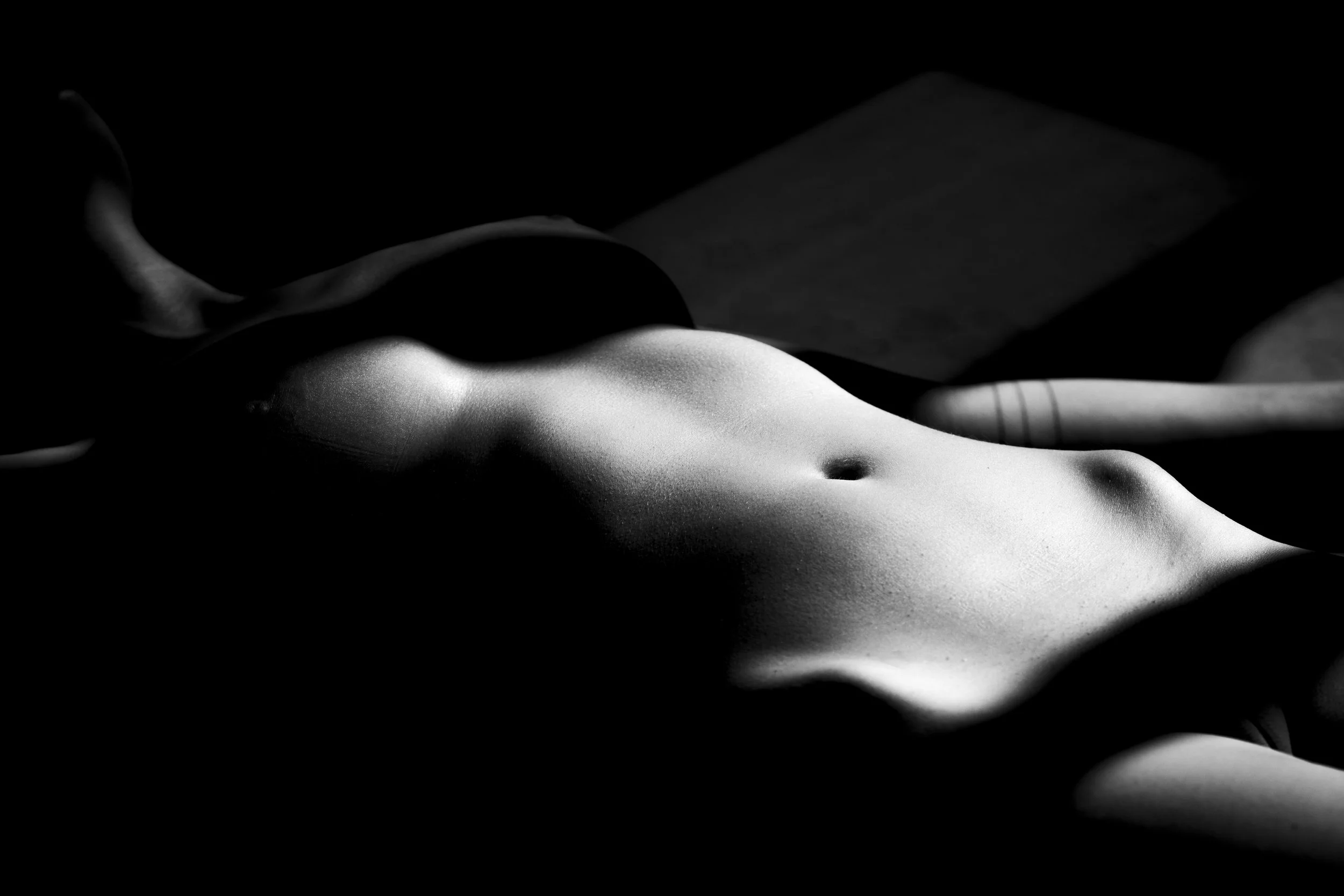 A black and white photo of a nude human body lying down with the right arm extended, emphasizing the curves and contours of the torso, with soft lighting creating highlights and shadows.