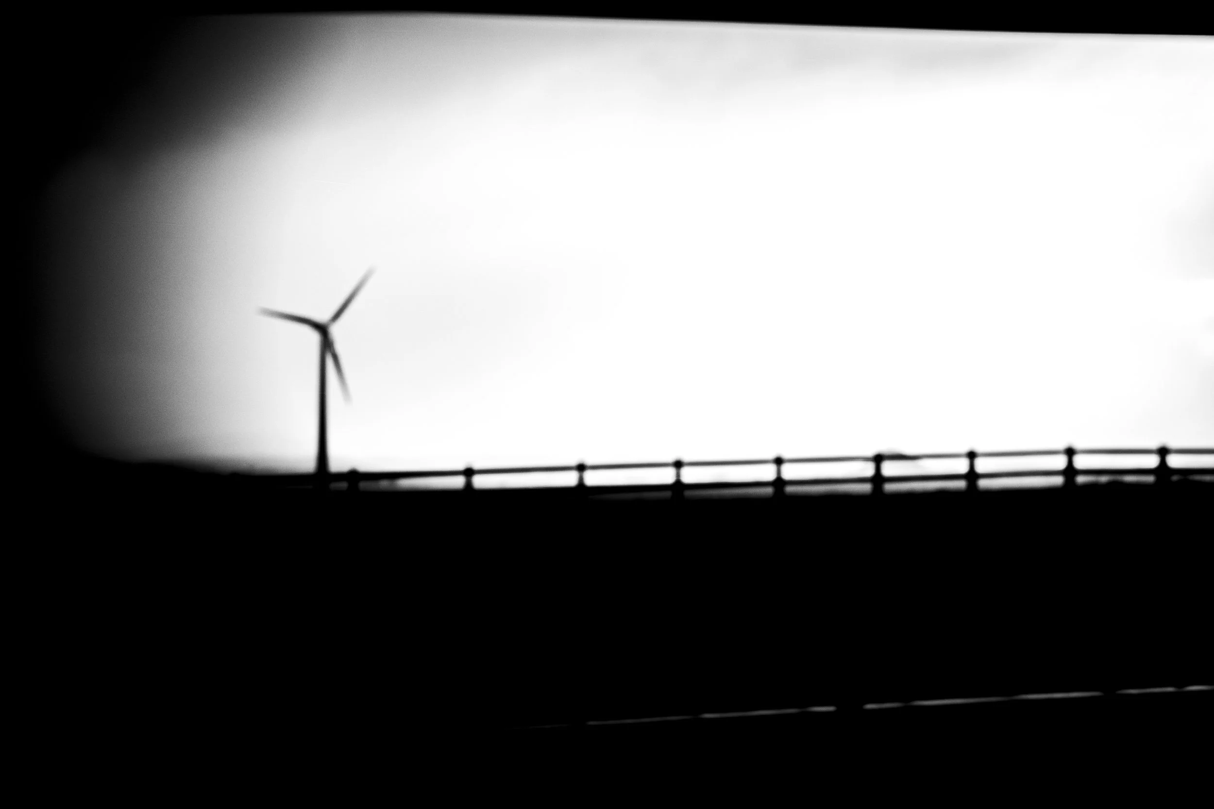 Black and white photo of a wind turbine, partially visible, alongside a roadway with a chain-link fence in the foreground.