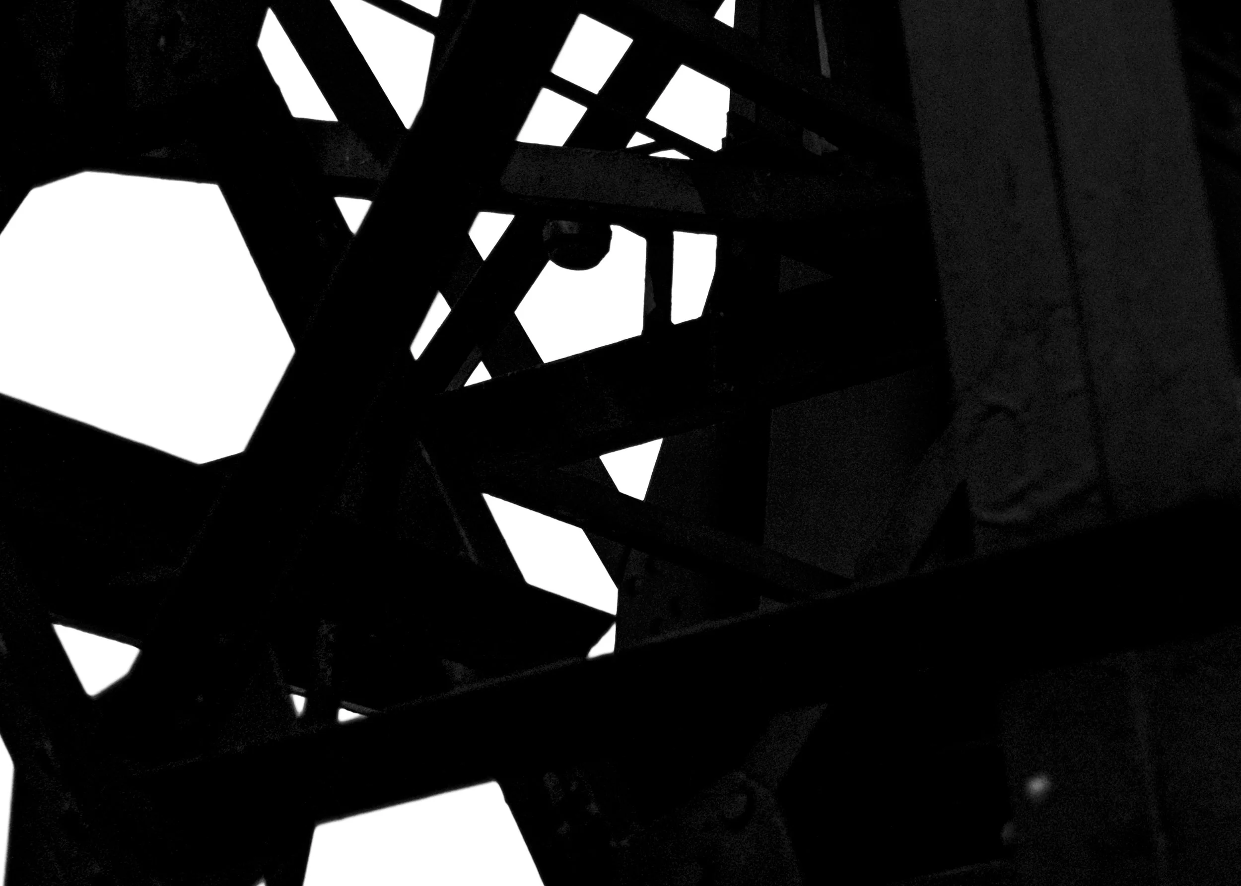 Black and white photograph of a geometric abstract sculpture with intersecting angular shapes and open spaces, viewed from below against a light background.