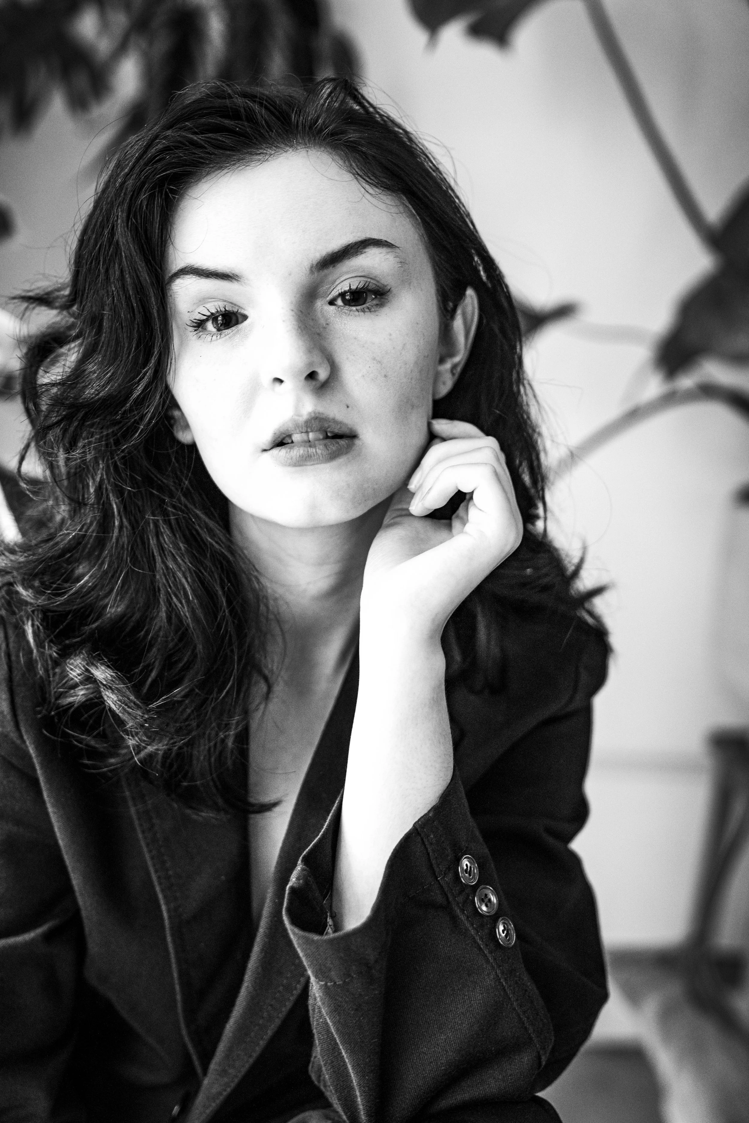 A black and white portrait of a young woman with long wavy hair, sitting with her hand resting on her chin, wearing a dark jacket with buttons.