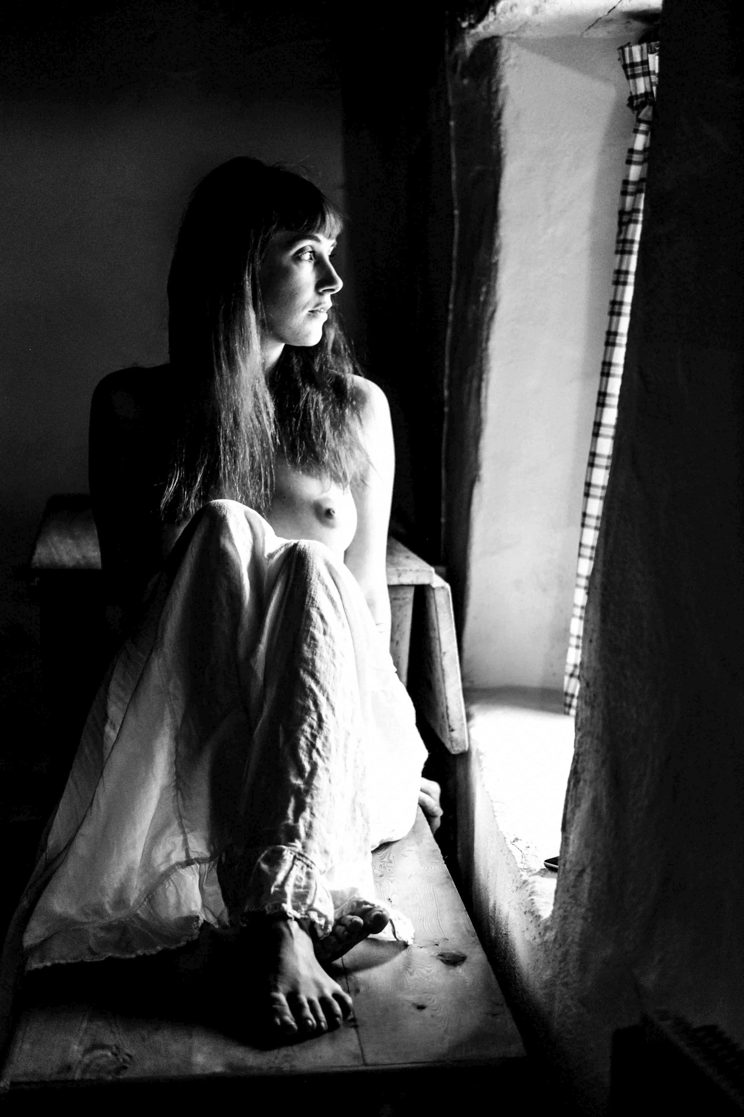 A woman with long hair sitting on the floor, looking out a window with plaid curtains, with sunlight illuminating her face in a black and white photograph.