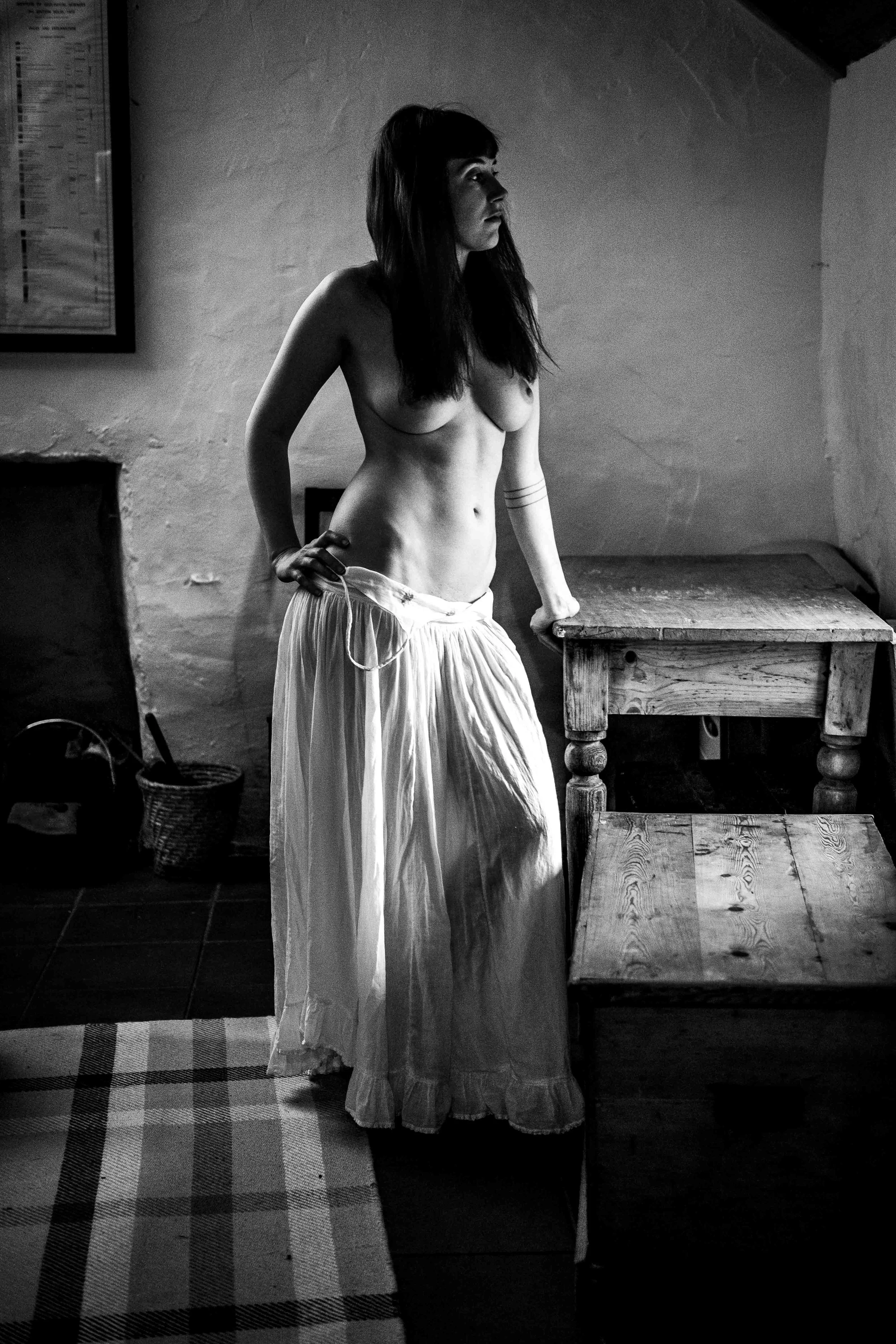 A black-and-white photo of a woman with dark hair standing in a room, topless, wearing a long, flowing skirt. She is looking to the right, with her left hand on her hip and her right hand resting on a wooden table. The room has a textured wall, a framed picture or chart, a woven basket on the floor, and a checkered rug.